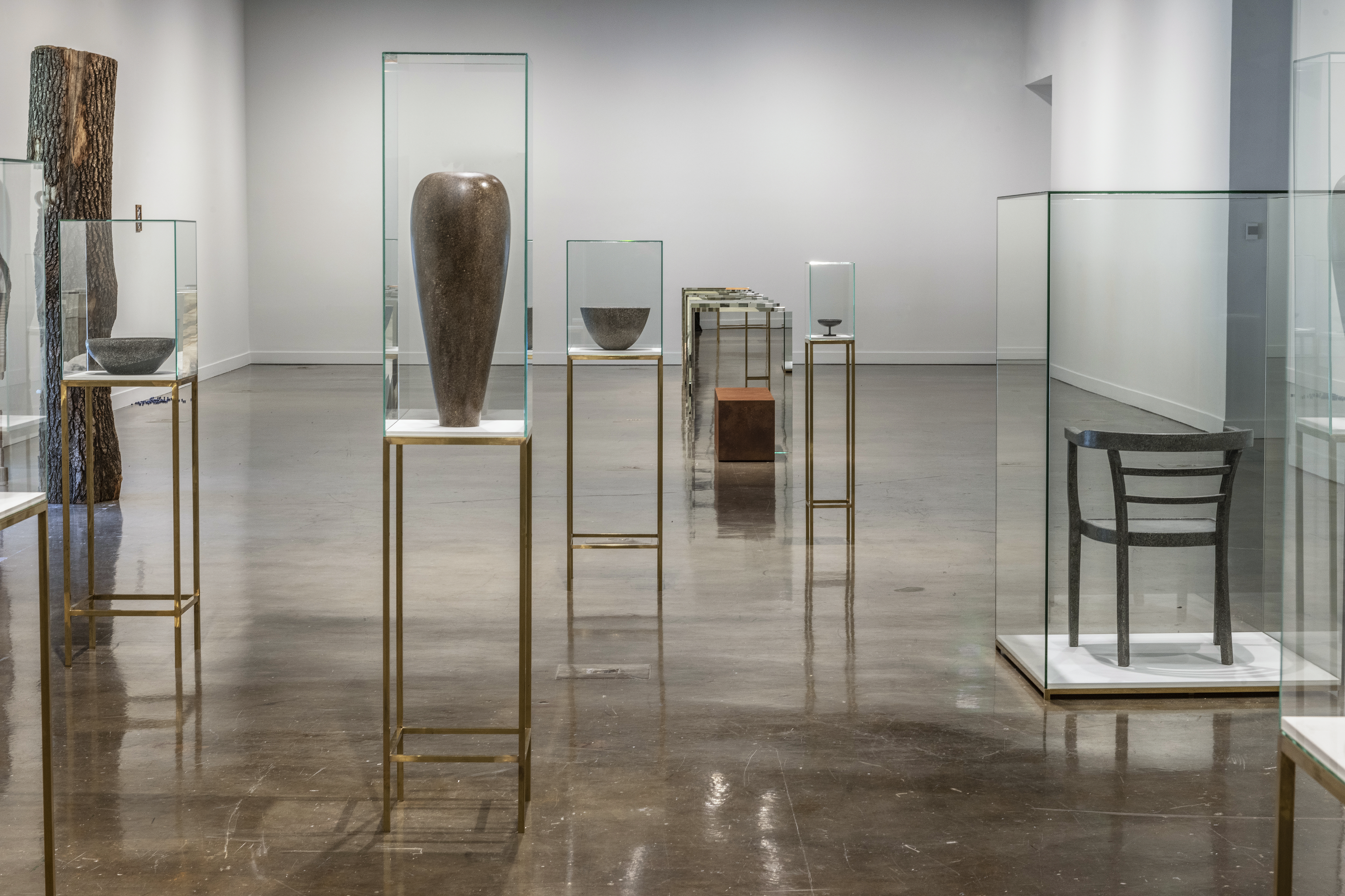 Vases and chairs seemingly made of concrete and metal sit atop bronze pedestals inside a glass case. Int he background are other works from Kwade's exhibition.