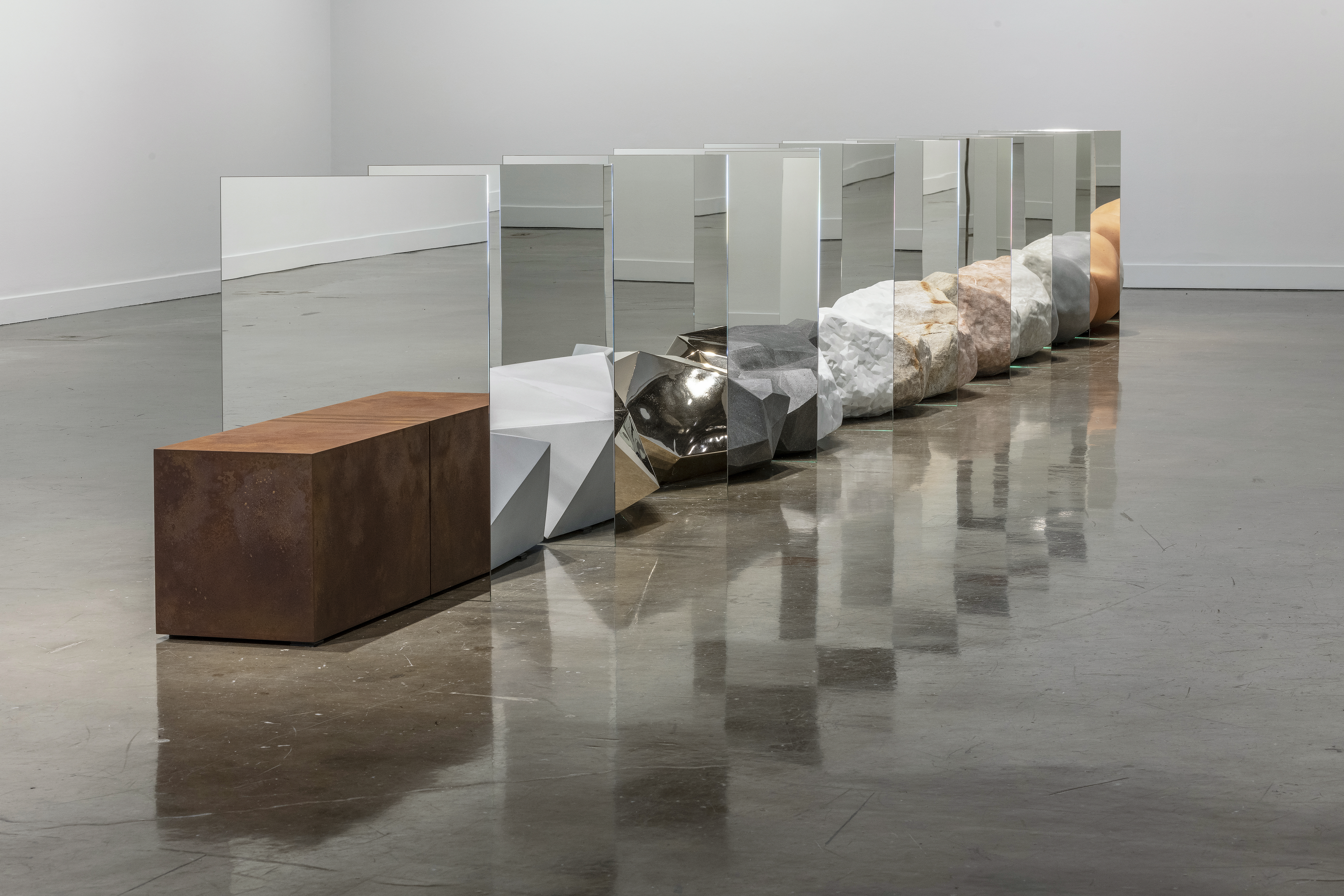 Diagonal view of objects resembling precious rocks are arraged in a straight row. Between each object is a mirror, creating the illusion that each rock is morphing into one another.