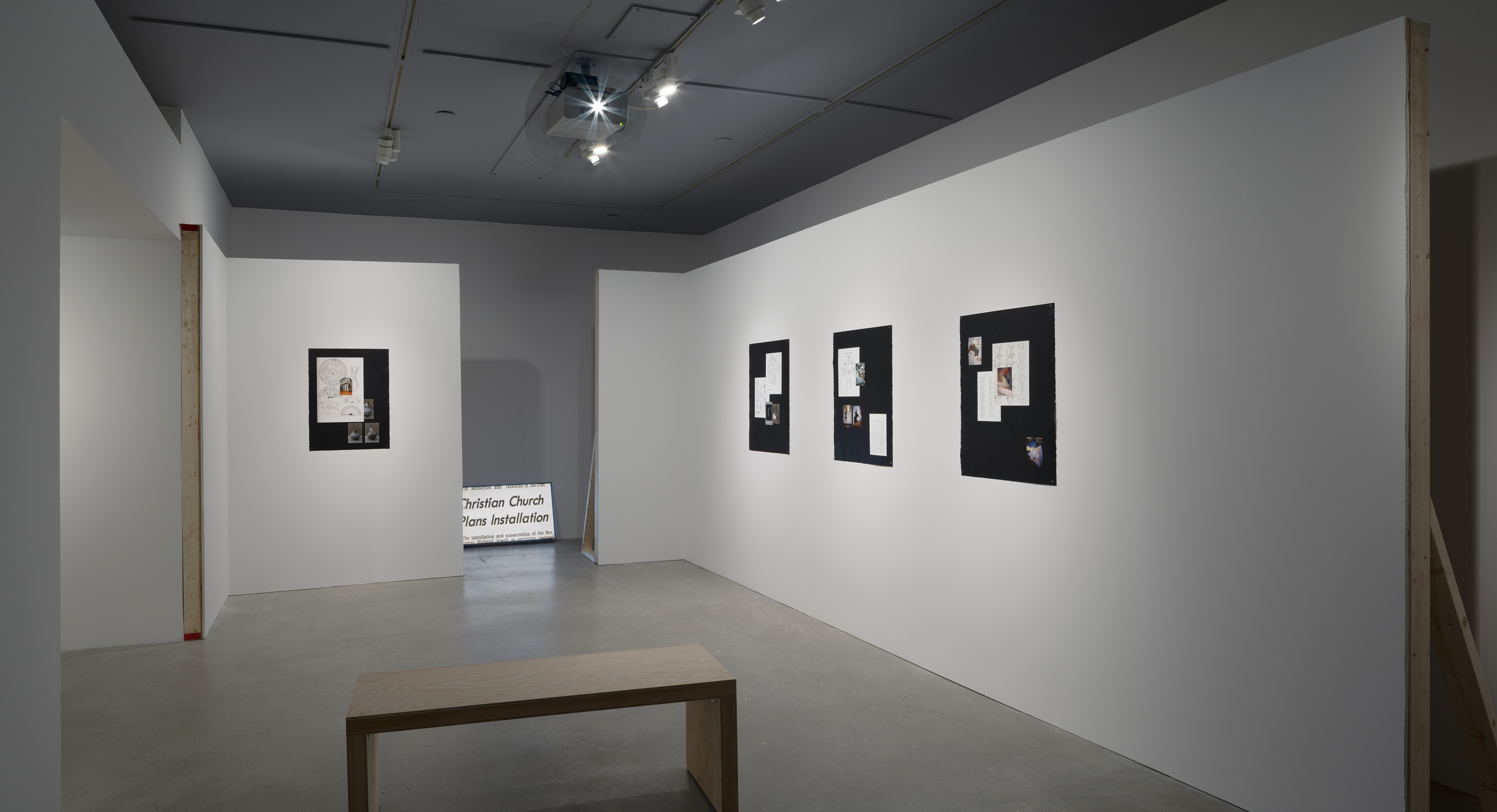 Wood bench in front of 4 paper works mounted on 2 constructed walls and a monitor on the floor between the 2 back walls