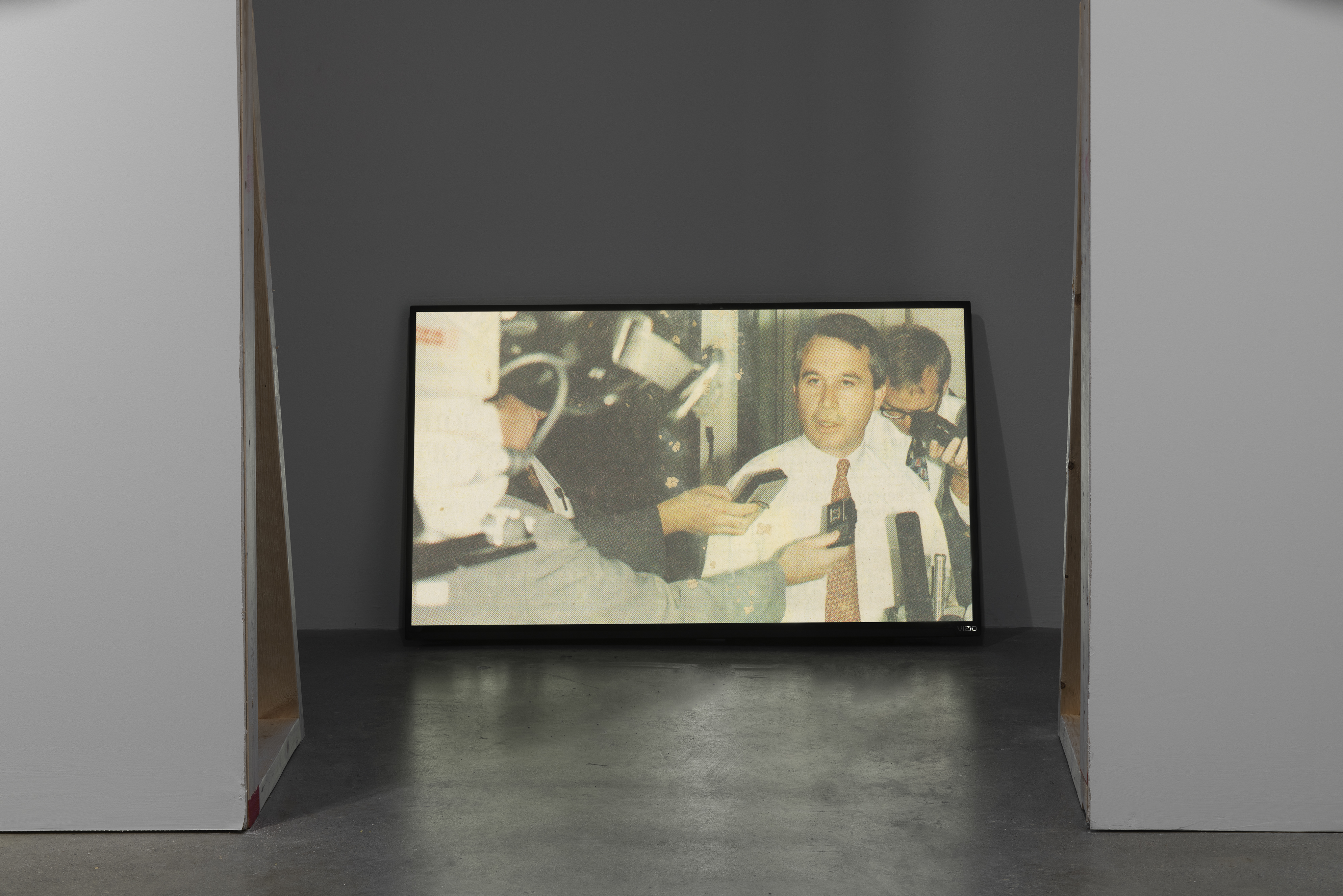 Seen from waist up, a man in shirt and tie speaks into tape recorders and mics in this still shown on a floor monitor.