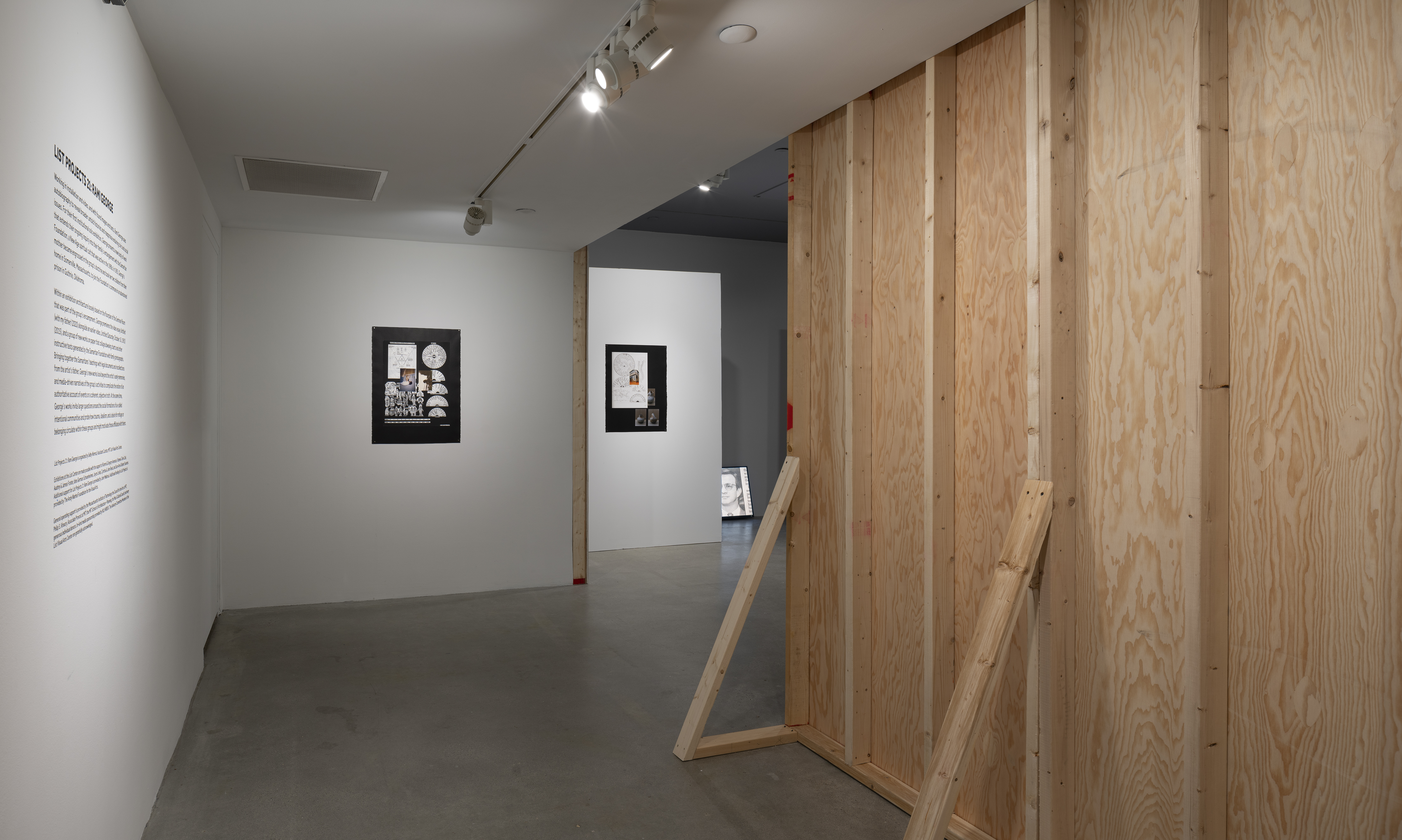 Exposed wood frame of the back of a wall, 2 paper works on black, wall text and a partial view of a monitor on the floor