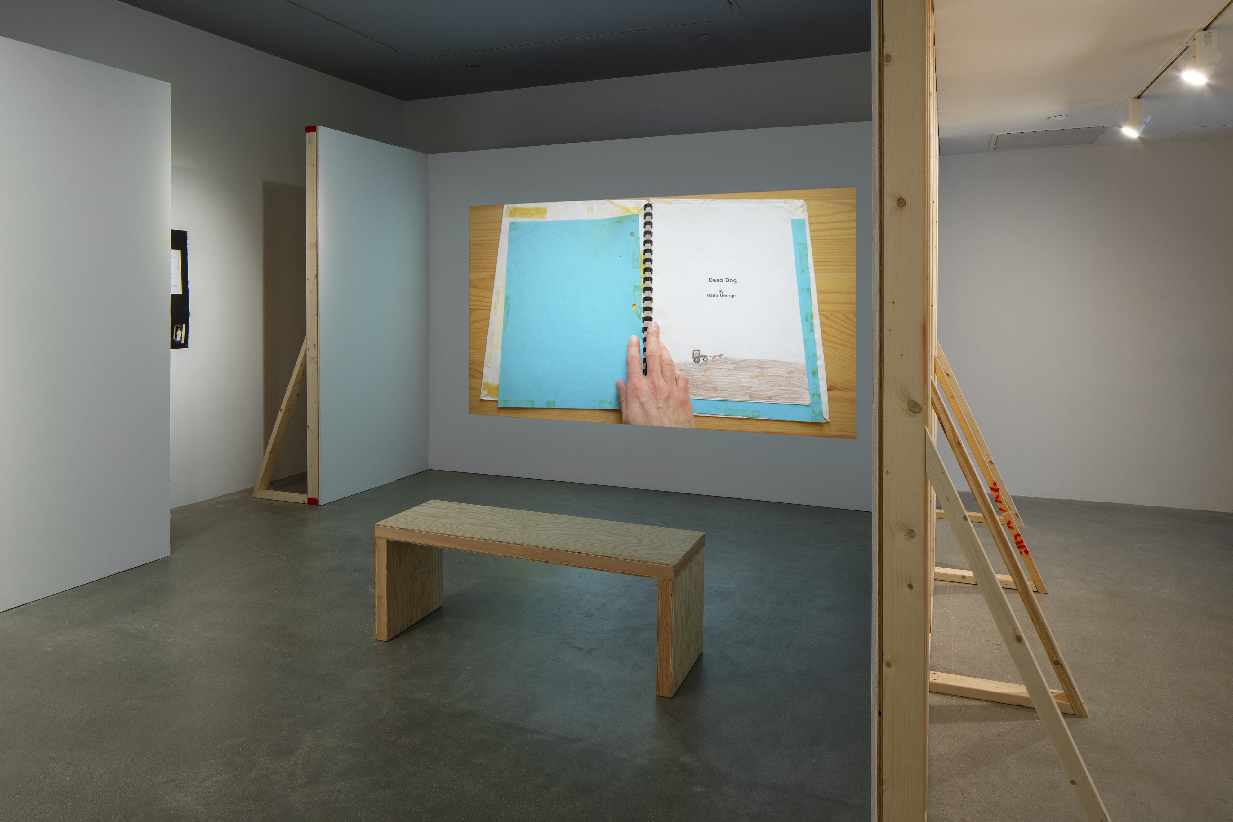 In a constructed alcove, a projected still of a child’s drawing titled “Dead Dog” in an old notebook with bright blue cover