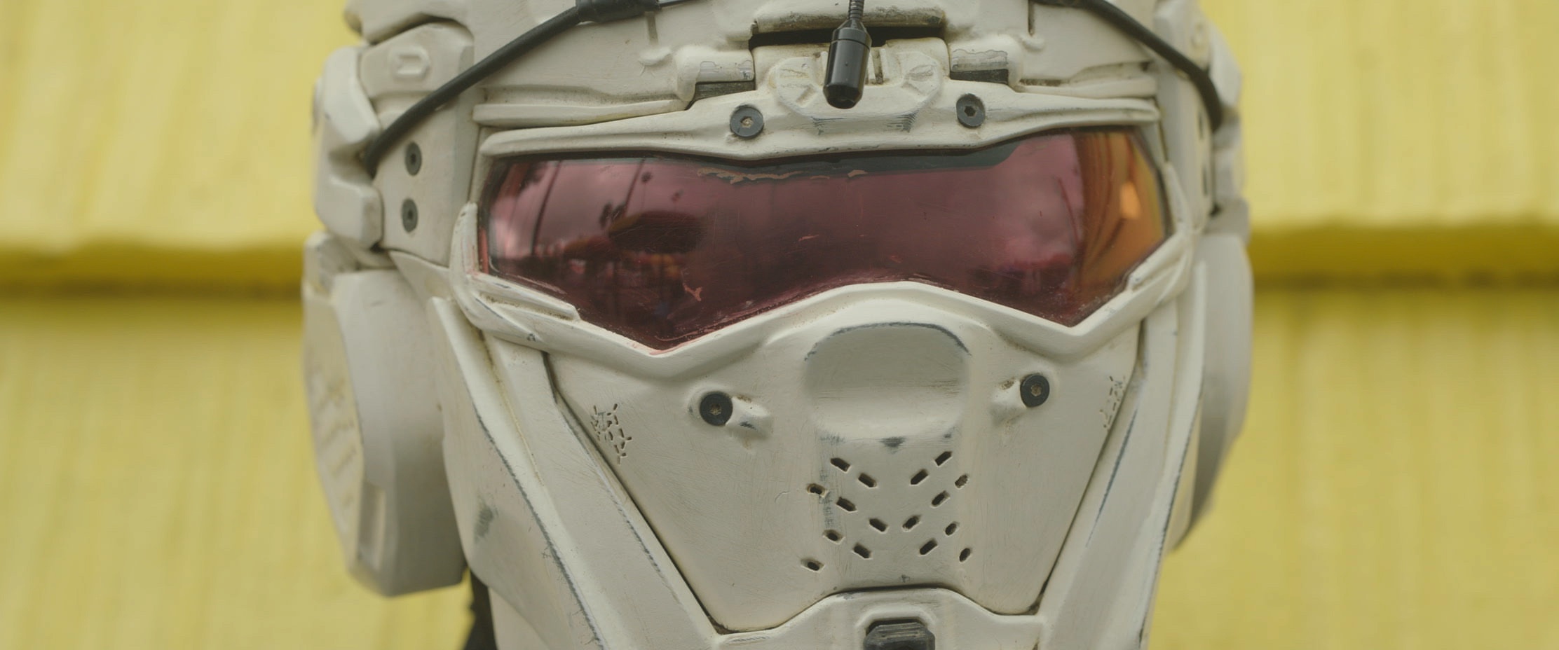 Extreme close up of a person wearing a futuristic storm-trooper-like helmet posing in front of bright yellow vinyl siding.