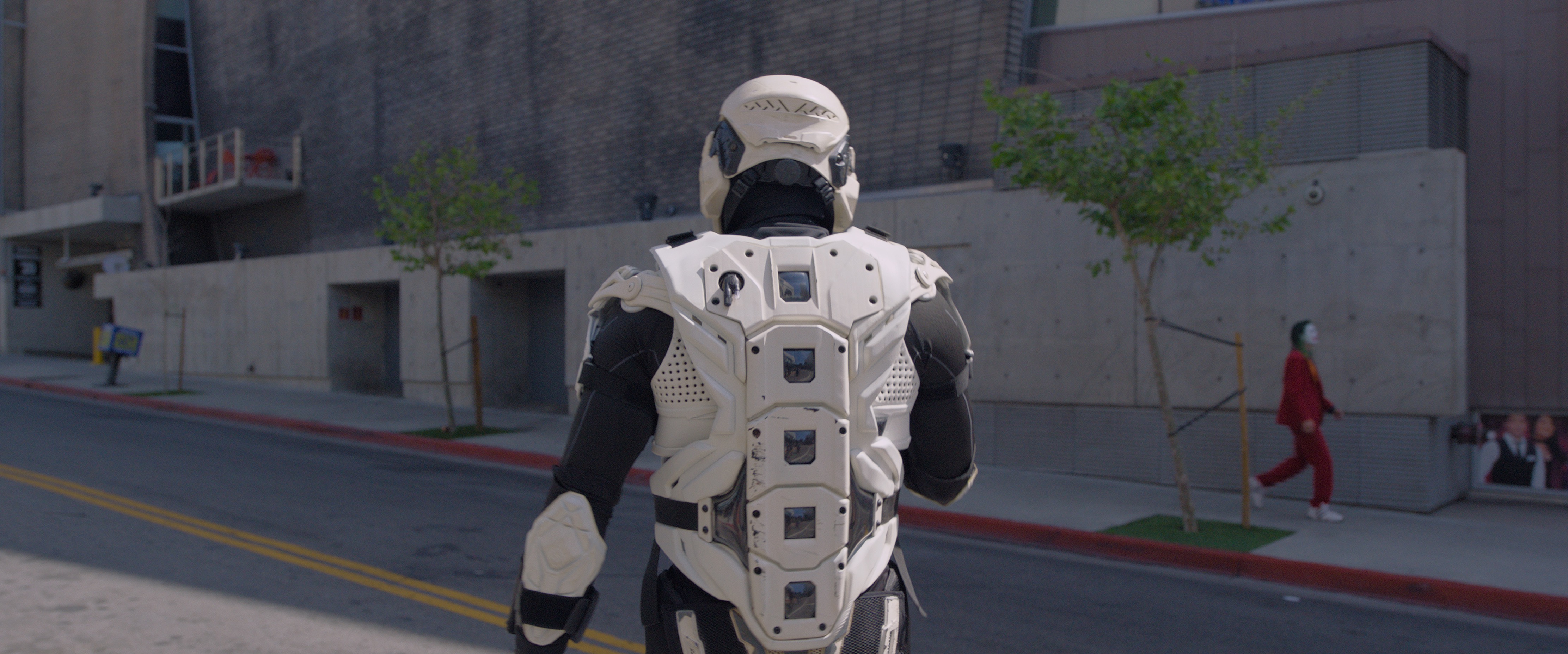 A figure in a black and white storm-trooper-like costume walks down the street with its back turned to the camera. A person in a red suit  green wig and Joker makeup walks on the distant opposing street.