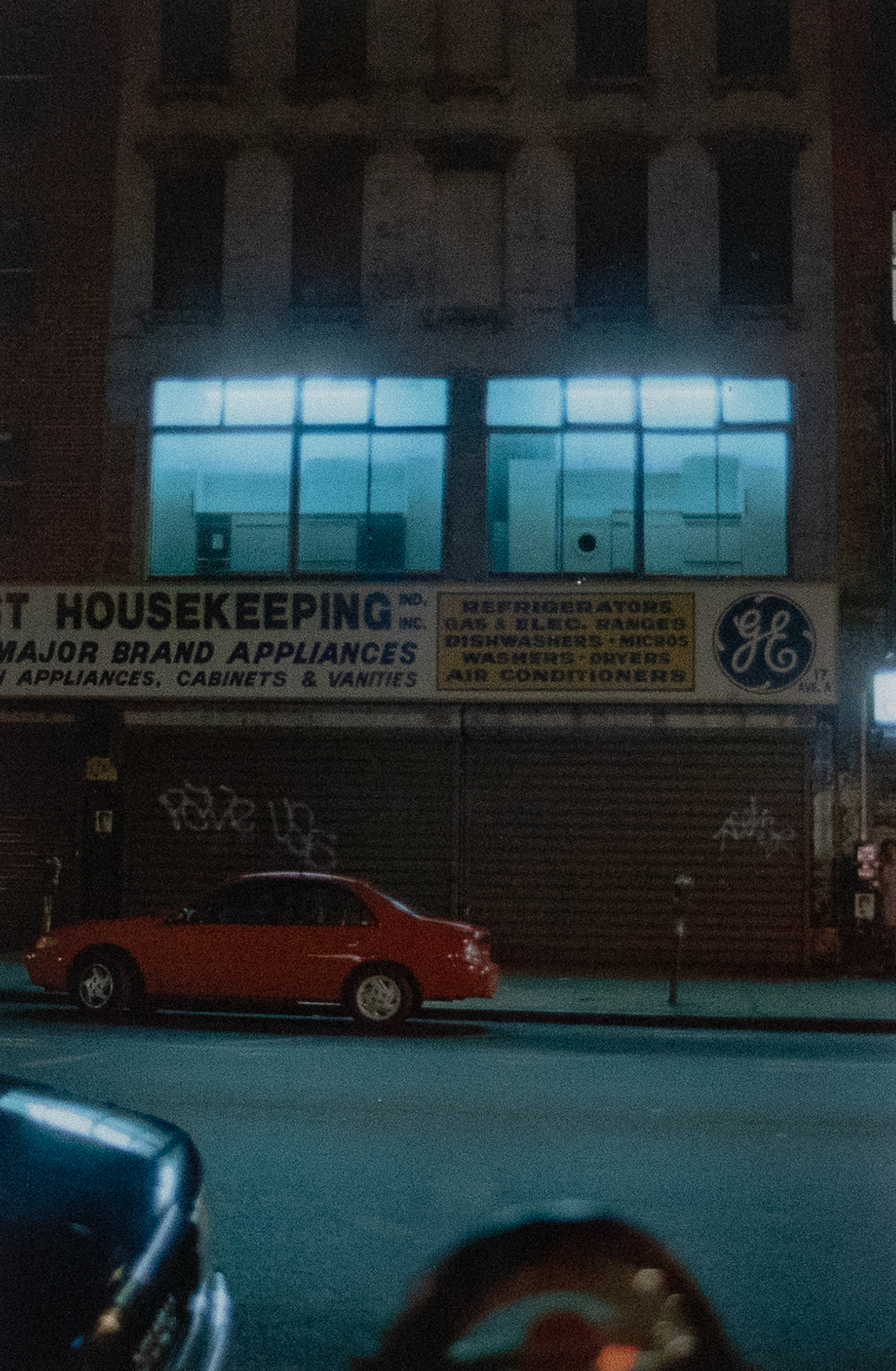 Night photograph of a storefront fro ma cross the street that reads &quot;Housekeeping&quot; and has a red car parked out front.