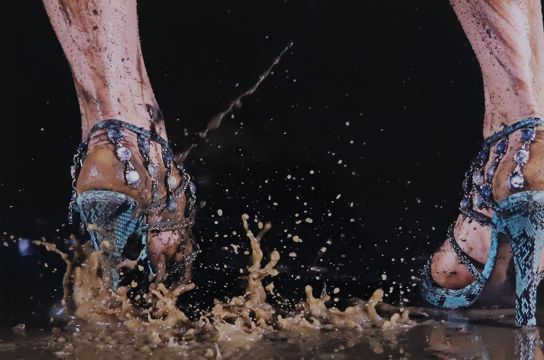 Close up photograph of a person wearing blue heels stepping in a brown puddle, making the water splatter all over.