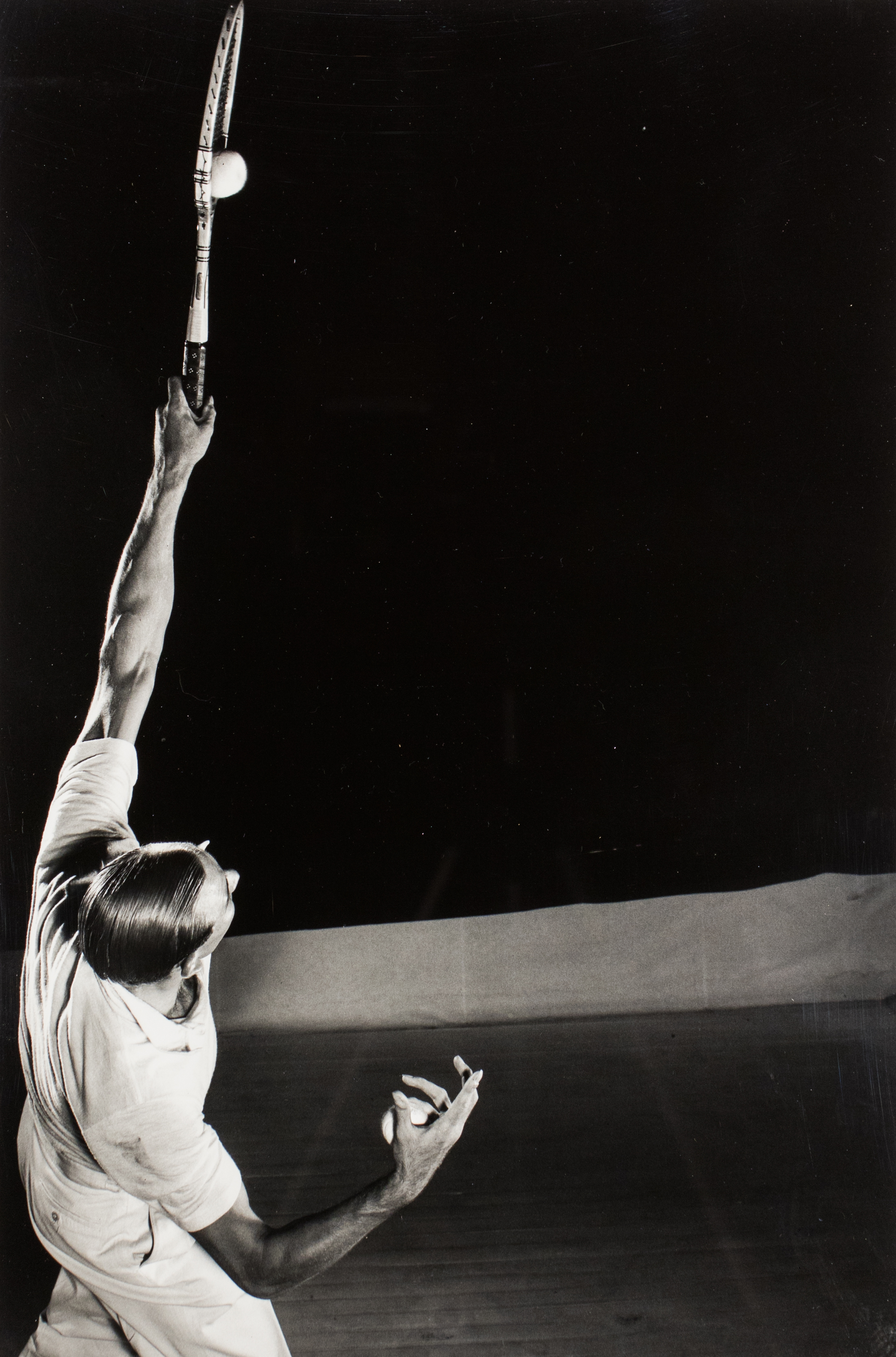 Black and white photograph of a man striking a tennis ball with his racket above his head.