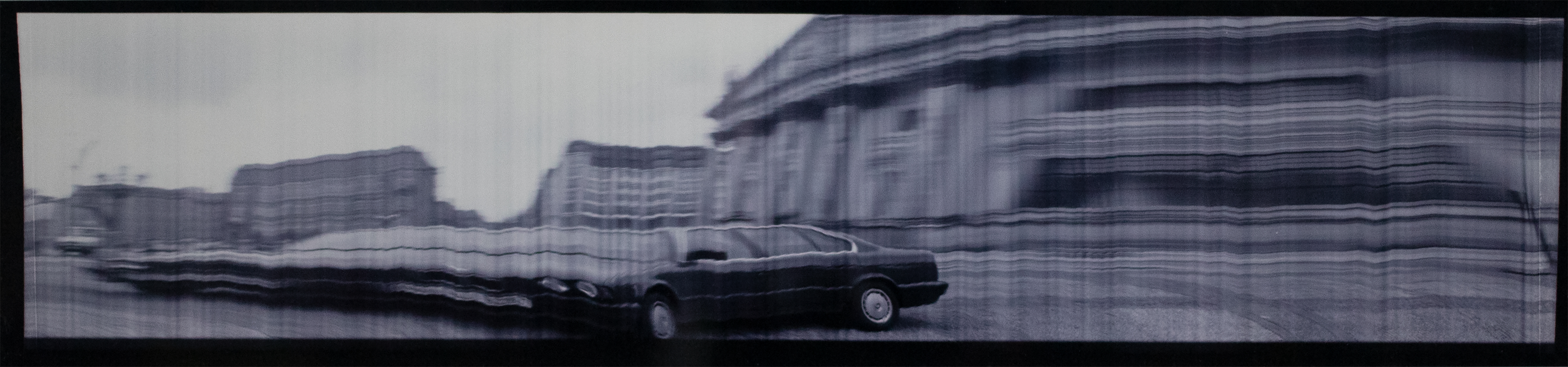 Photograph of car stretches out to the left of the frame with buildings in the background.