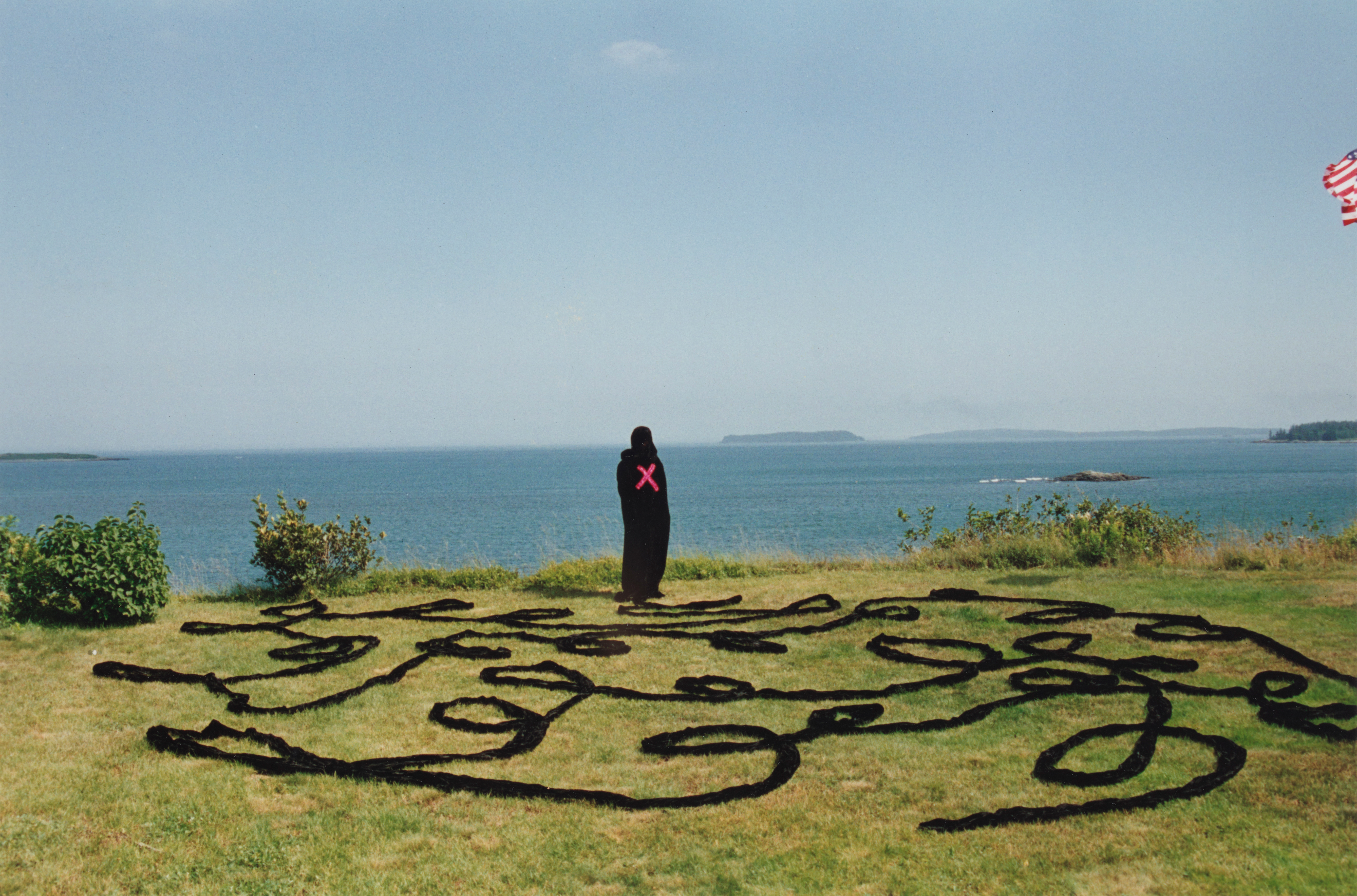 A person wearing all black with a large red X on their back stands on a grassy cliff that overlooks the ocean. The grass has a series of dark ropes in loops.