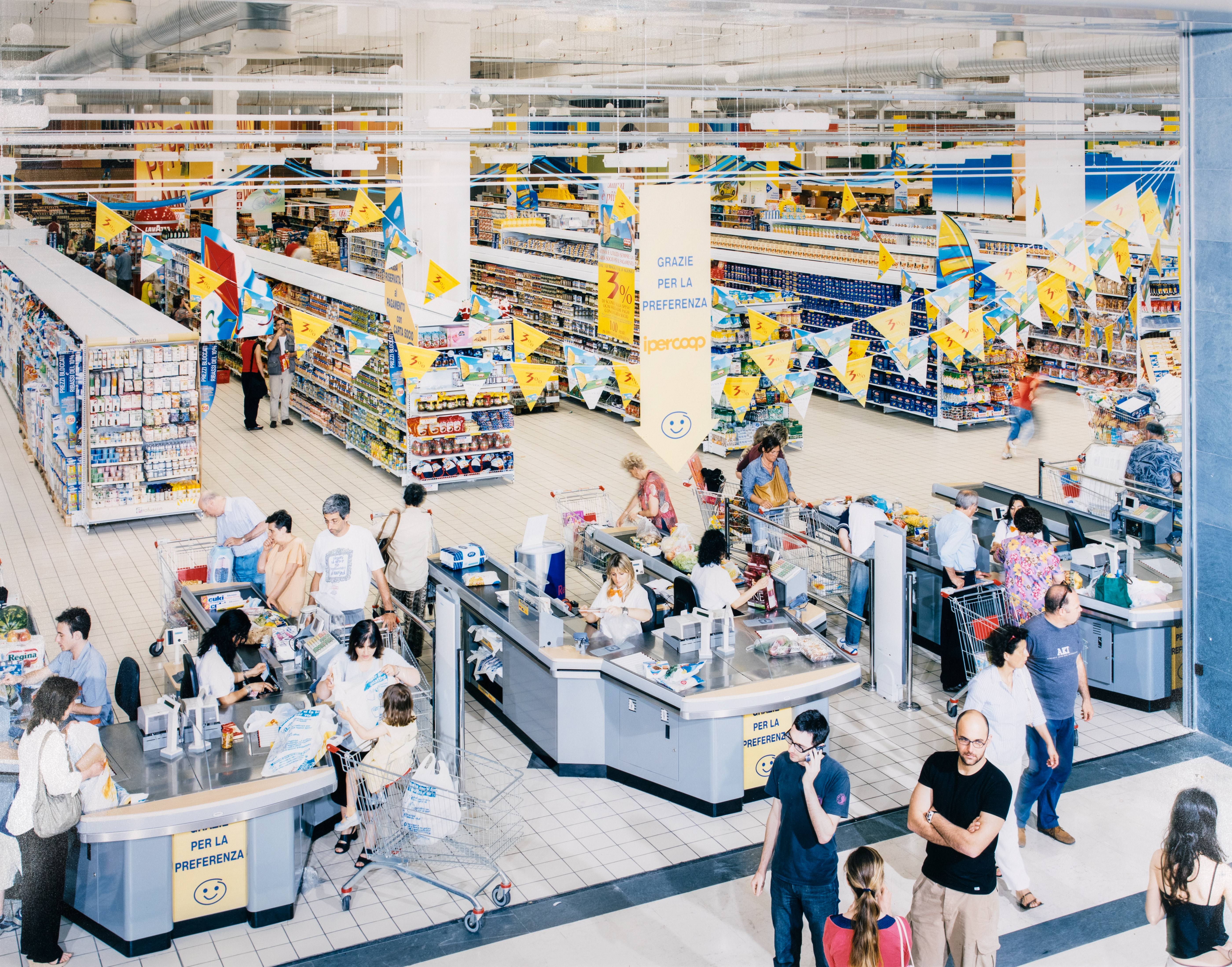 Photograph taken from up high busy checkout aisles at a supermarket featuring yellow flags hanging on the ceiling and the store aisles in the background.