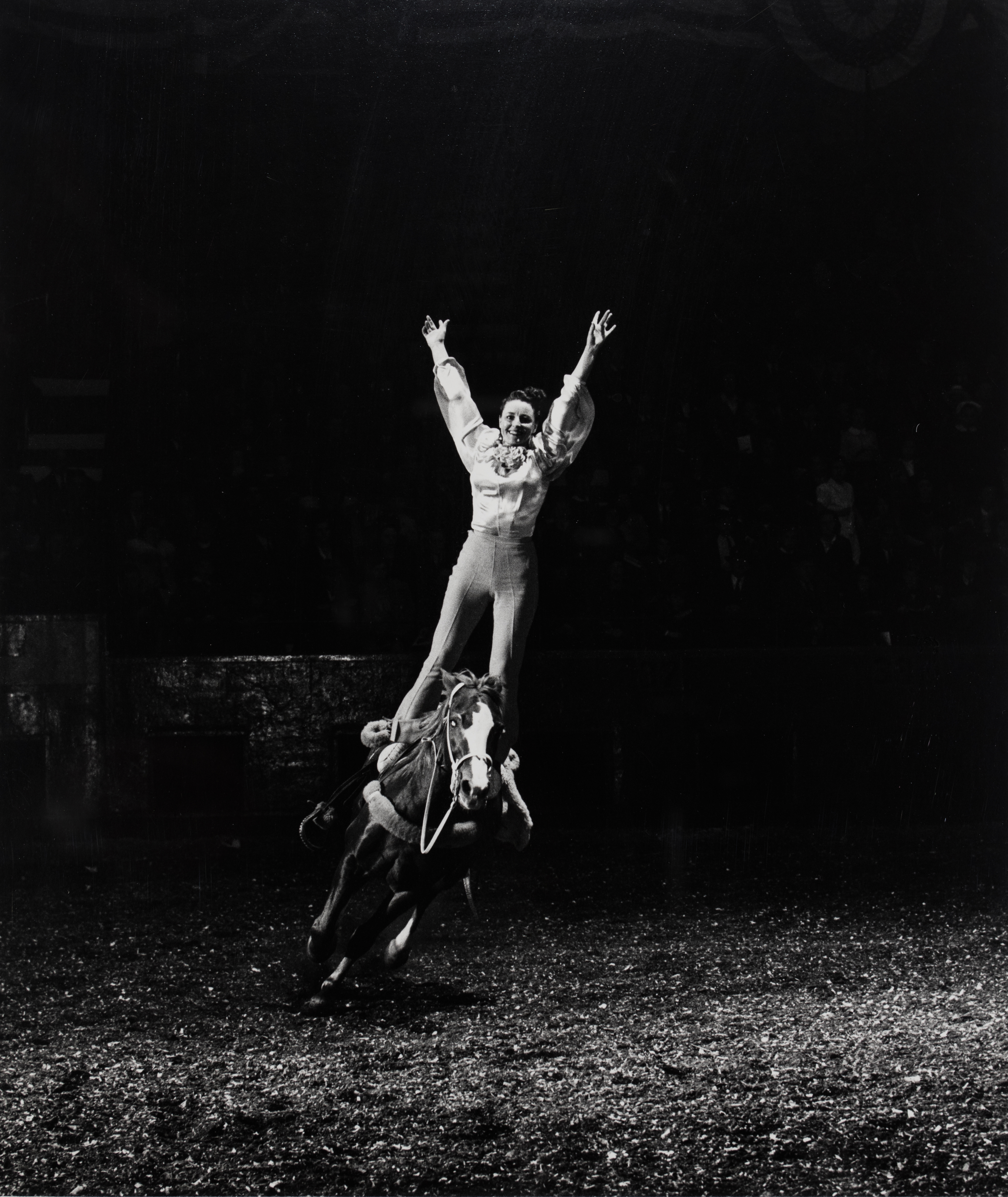 Black and white photograph of a girl standing straight up on a horse with her hands in the air.