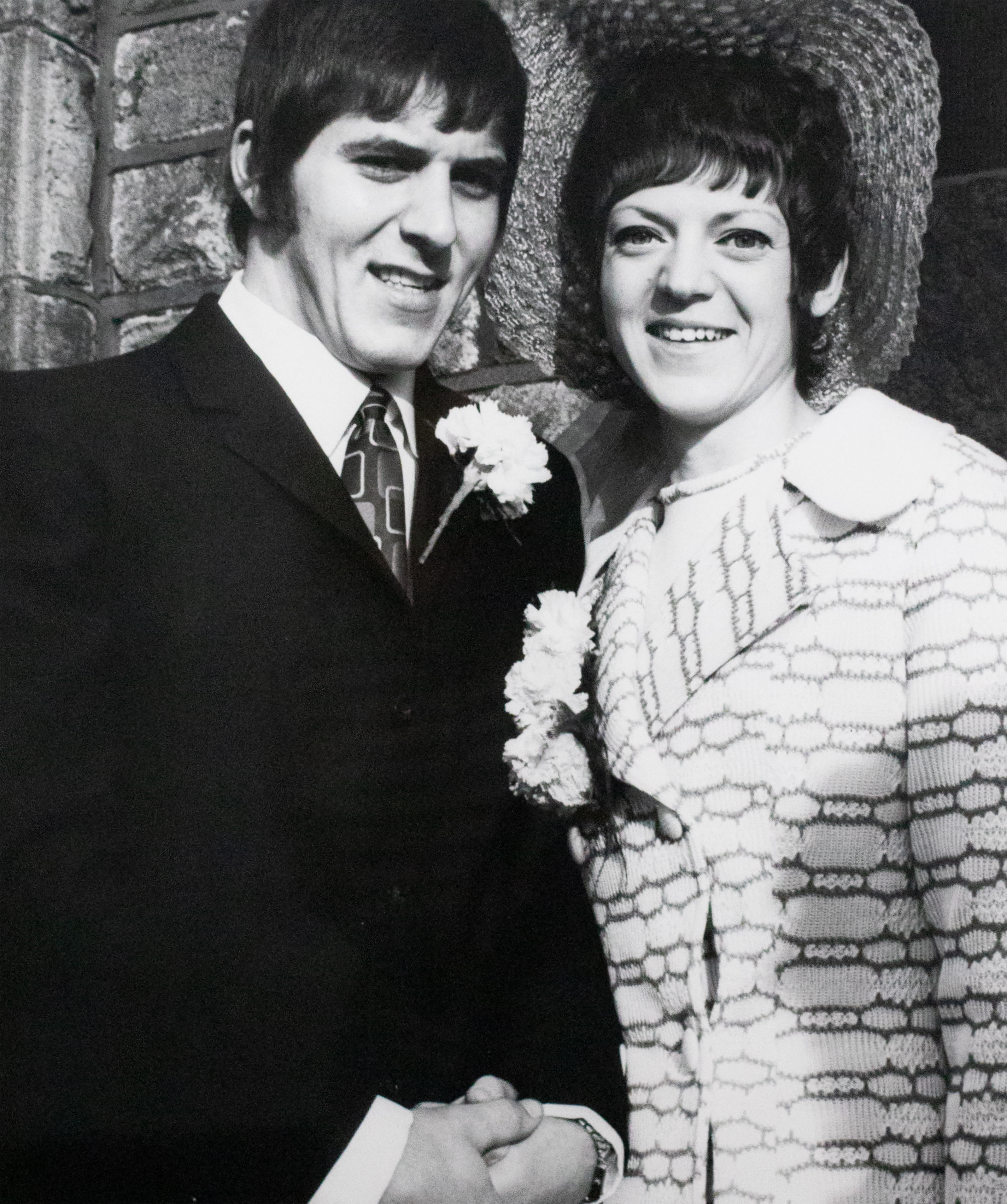 A man in a suit stands next to a woman in a dress with a fancy hat and they smile at the camera.