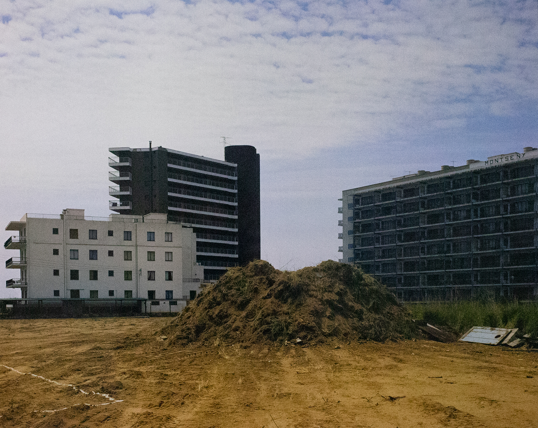 Photograph of a construction site with a large pile of dirt and three long buildings in the background.