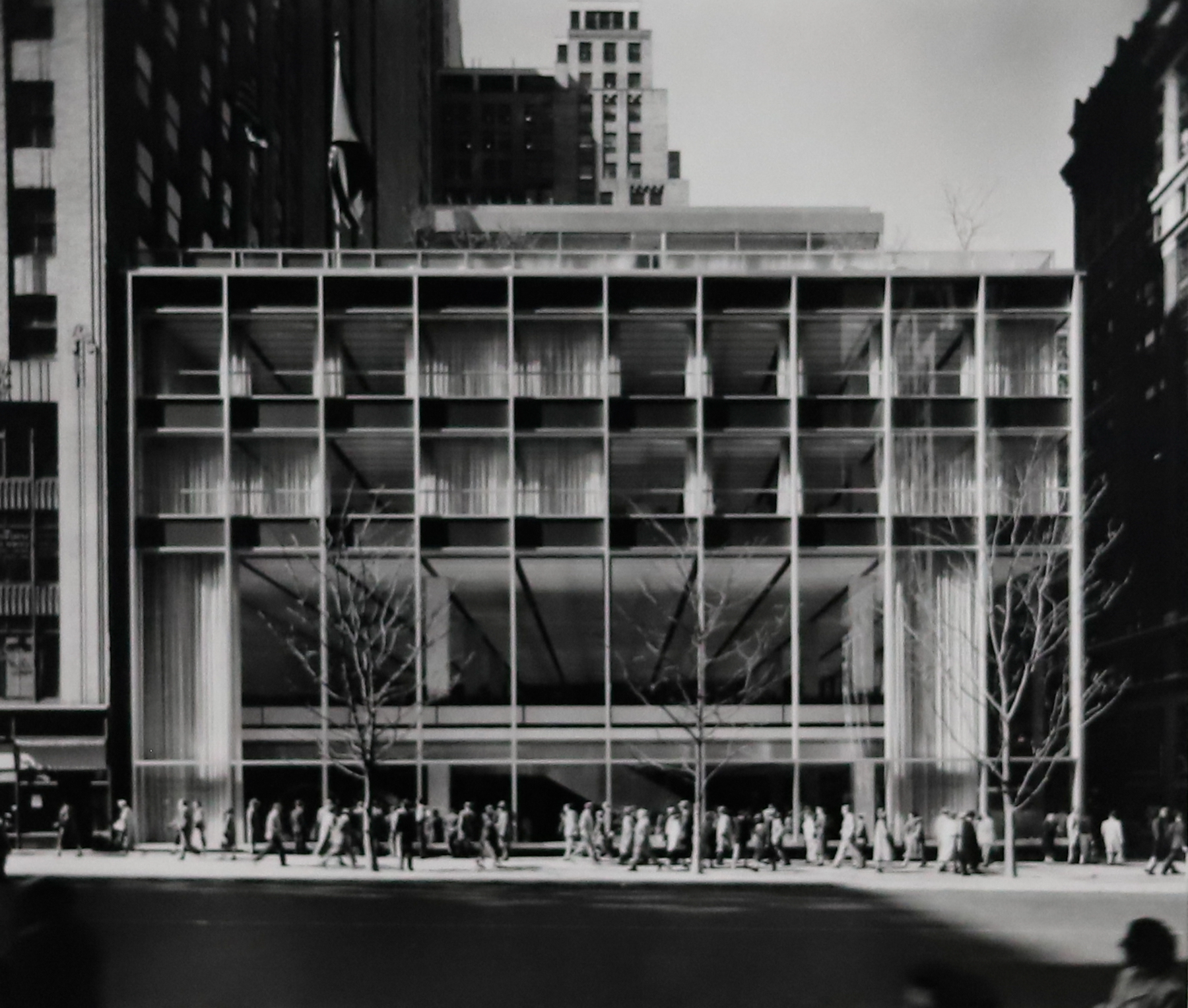 Black and white photograph of a building with tall paneled windows and a long, straight line of people walking out front.