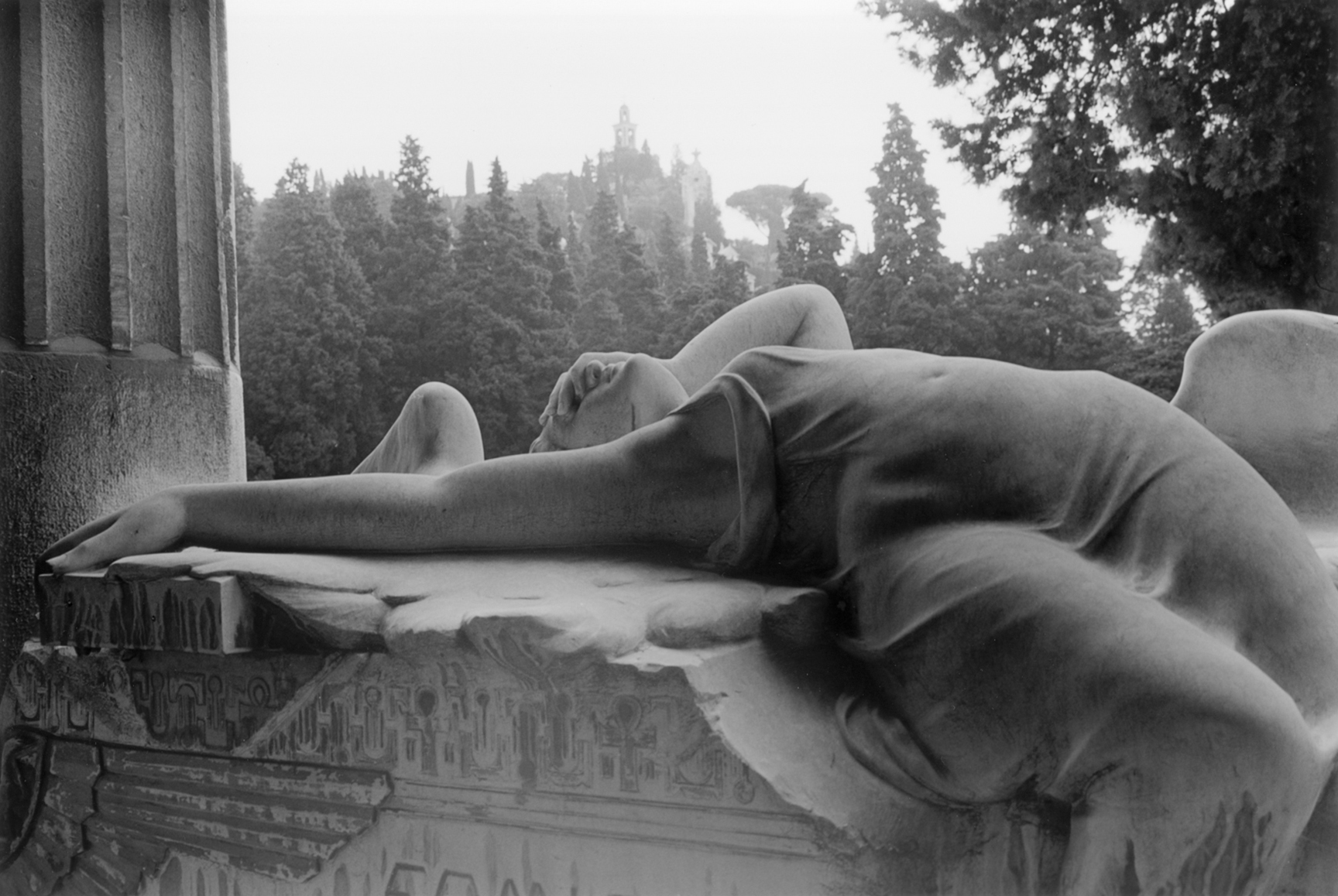 Black and white photograph of a woman lying on the edge of a balcony looking at the sky with trees in the background.