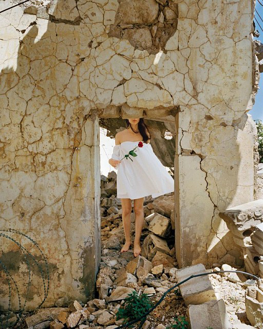 Photograph of a woman in a short white dress holding a rose standing under a doorway of crackling stone with her face cutoff at the top.