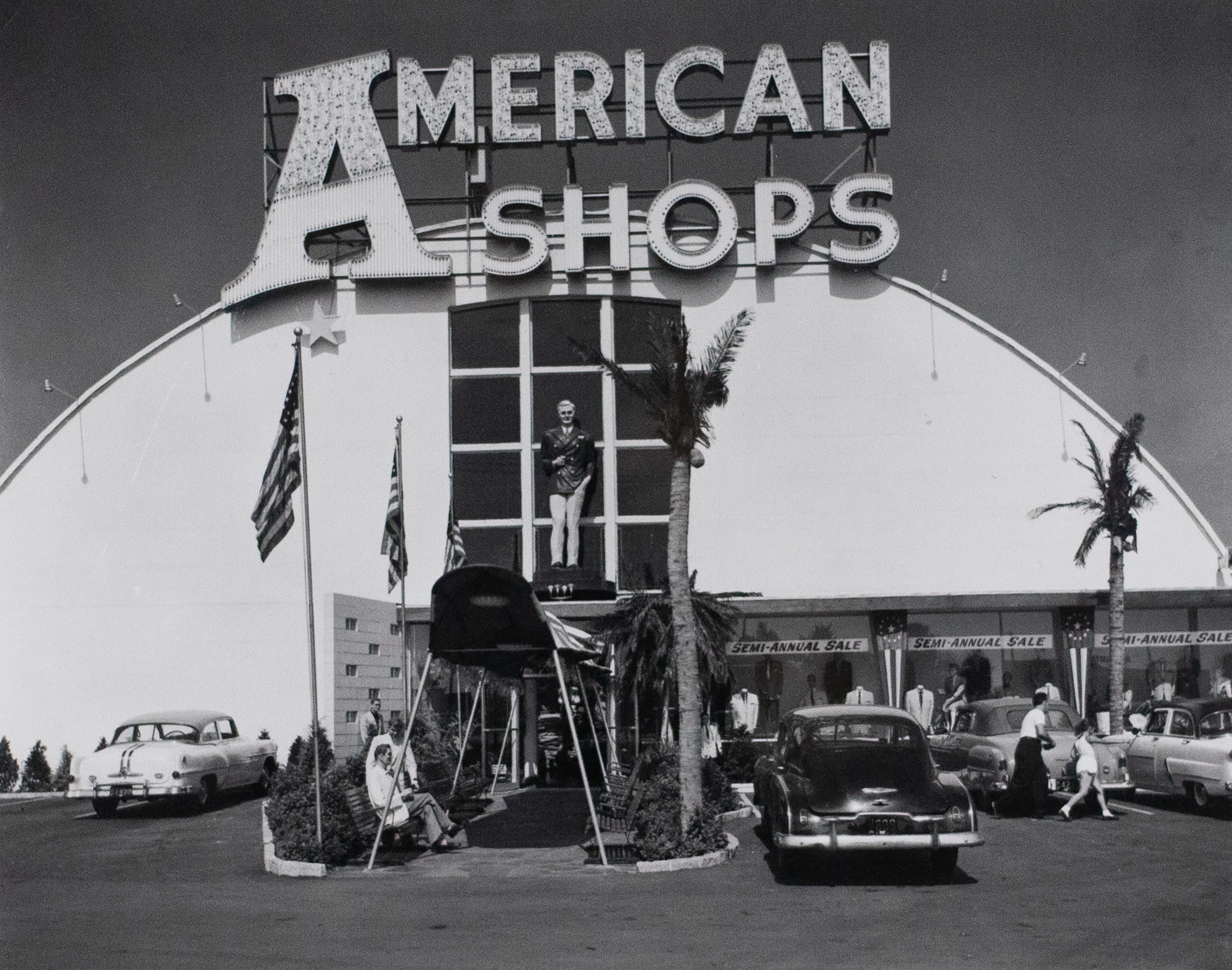 Photograph of a neon letter sign that reads &quot;American Shops&quot; in the parking lot with large shopping center behind.
