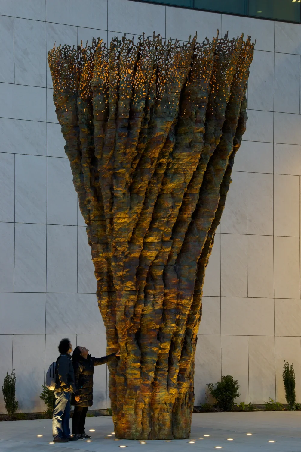 Bronze sculpture at night resembling a tree trunk with two people looking up at it on the sidewalk.