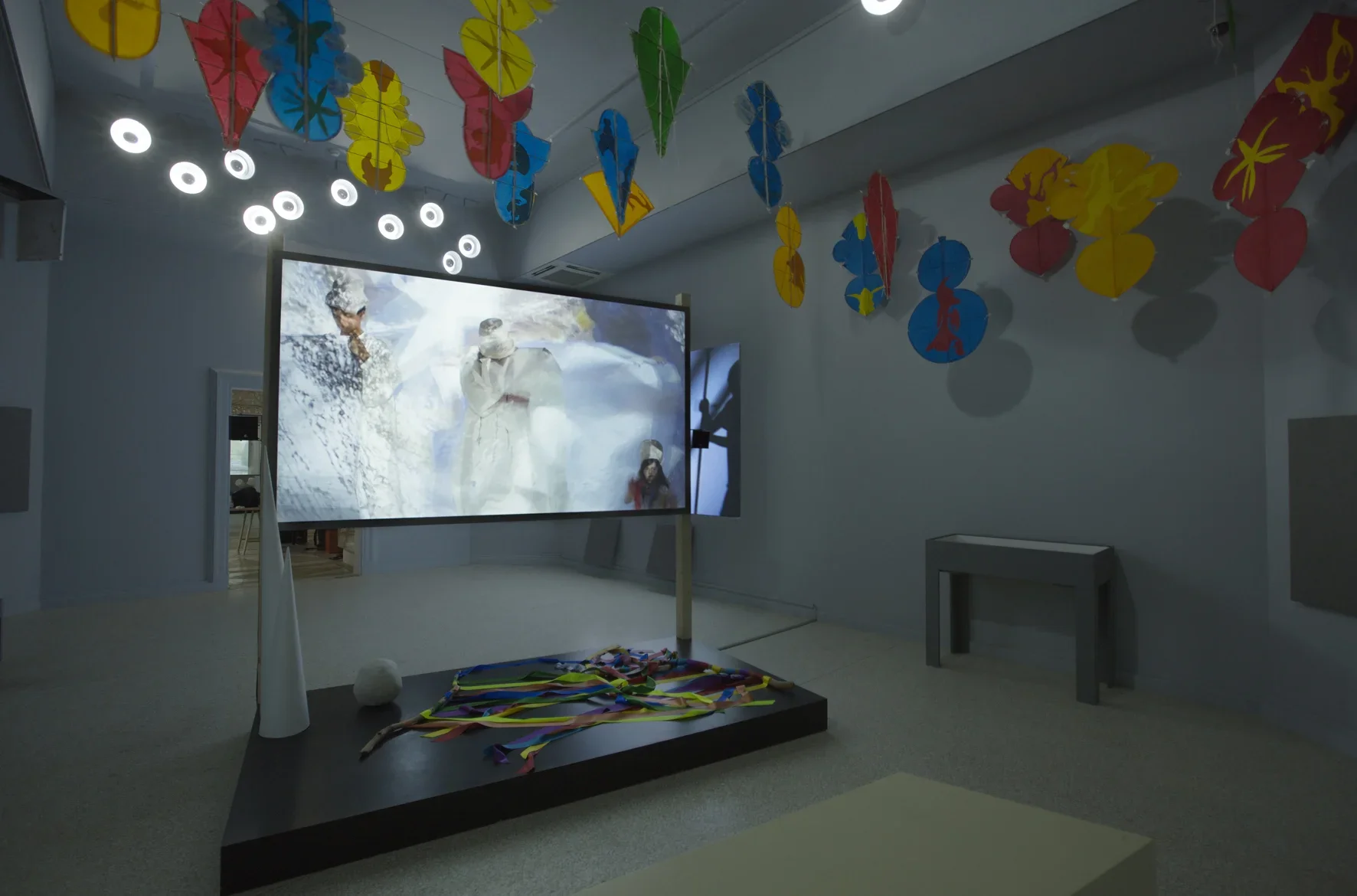 Installation view featuring a large screen in the center of the room playing a video and red, blue, and green shapes hanging from the ceiling.
