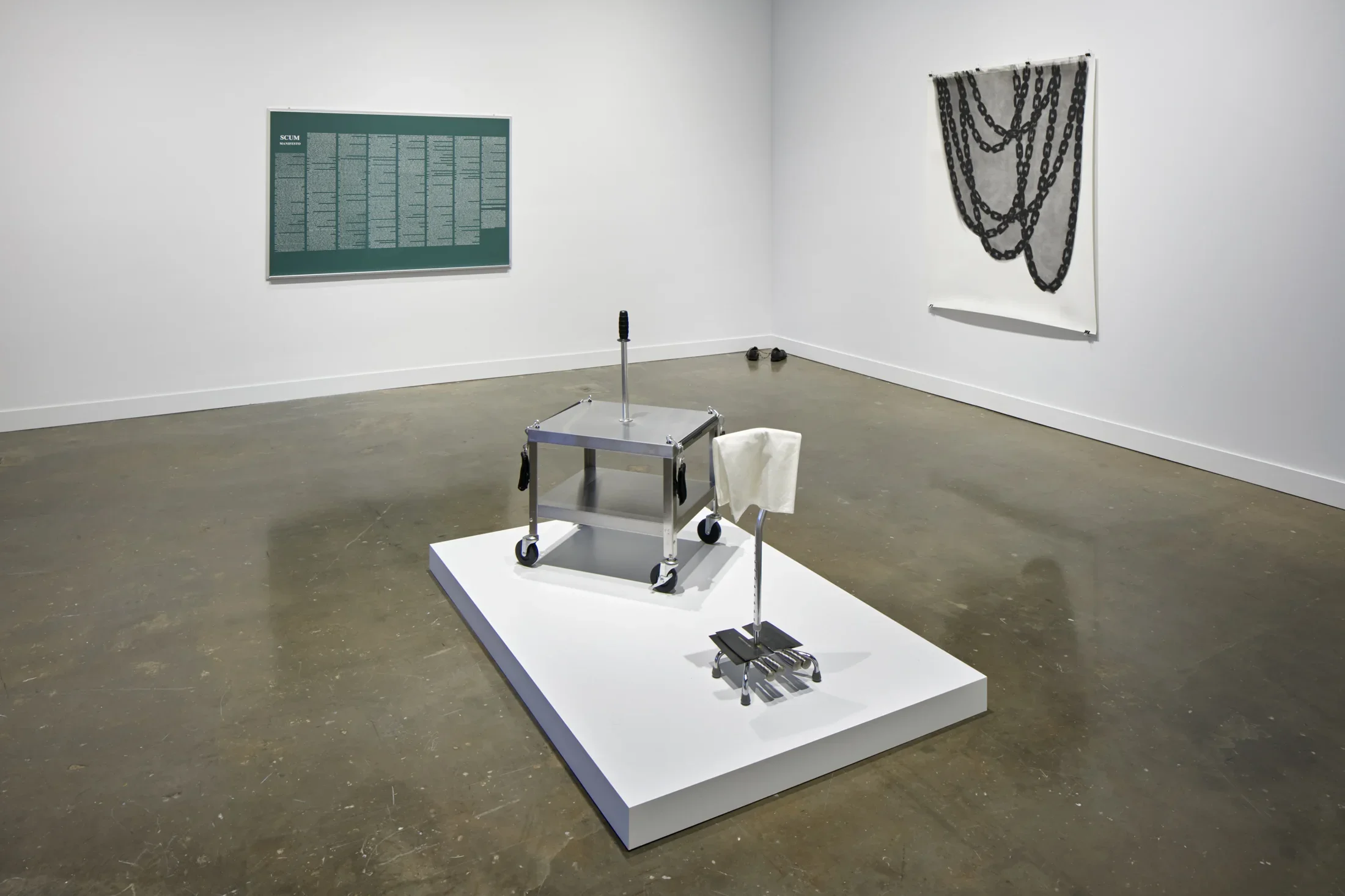 Installation view featuring a metal card on a podium in the center, a green print with white text on the wall, and a tapestry with chain like graphic on the right wall.