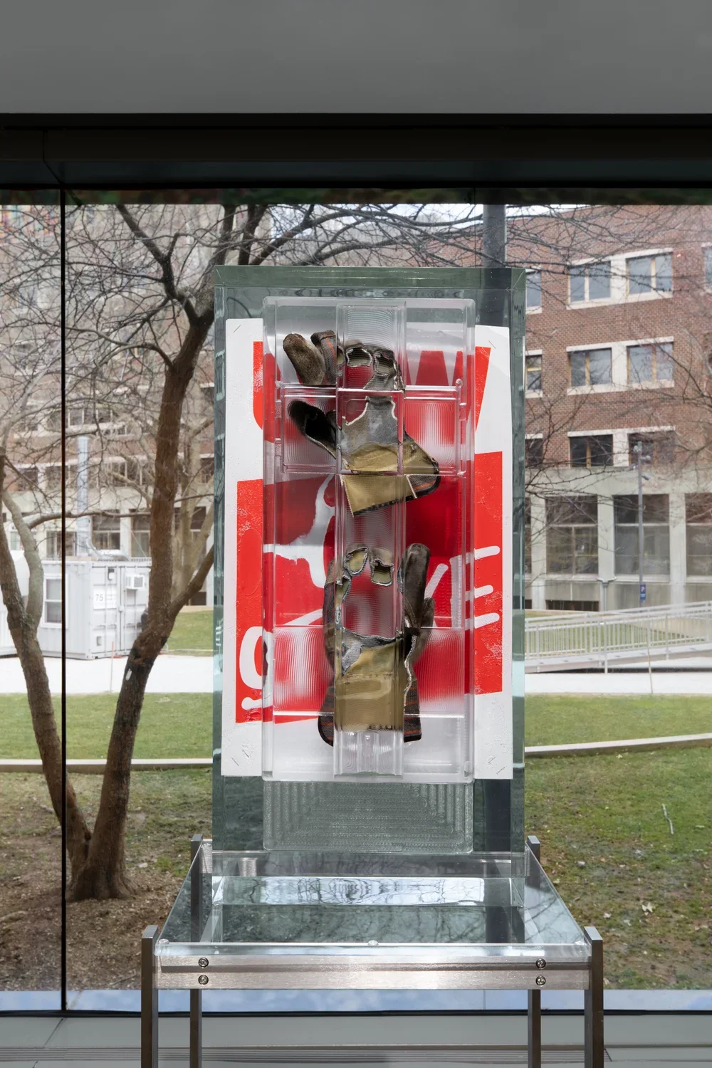 Installation view of a pair of work gloves and a red poster encased in a rectangular block of resin on a metal stand with a large window overlooking a courtyard in the background. 
