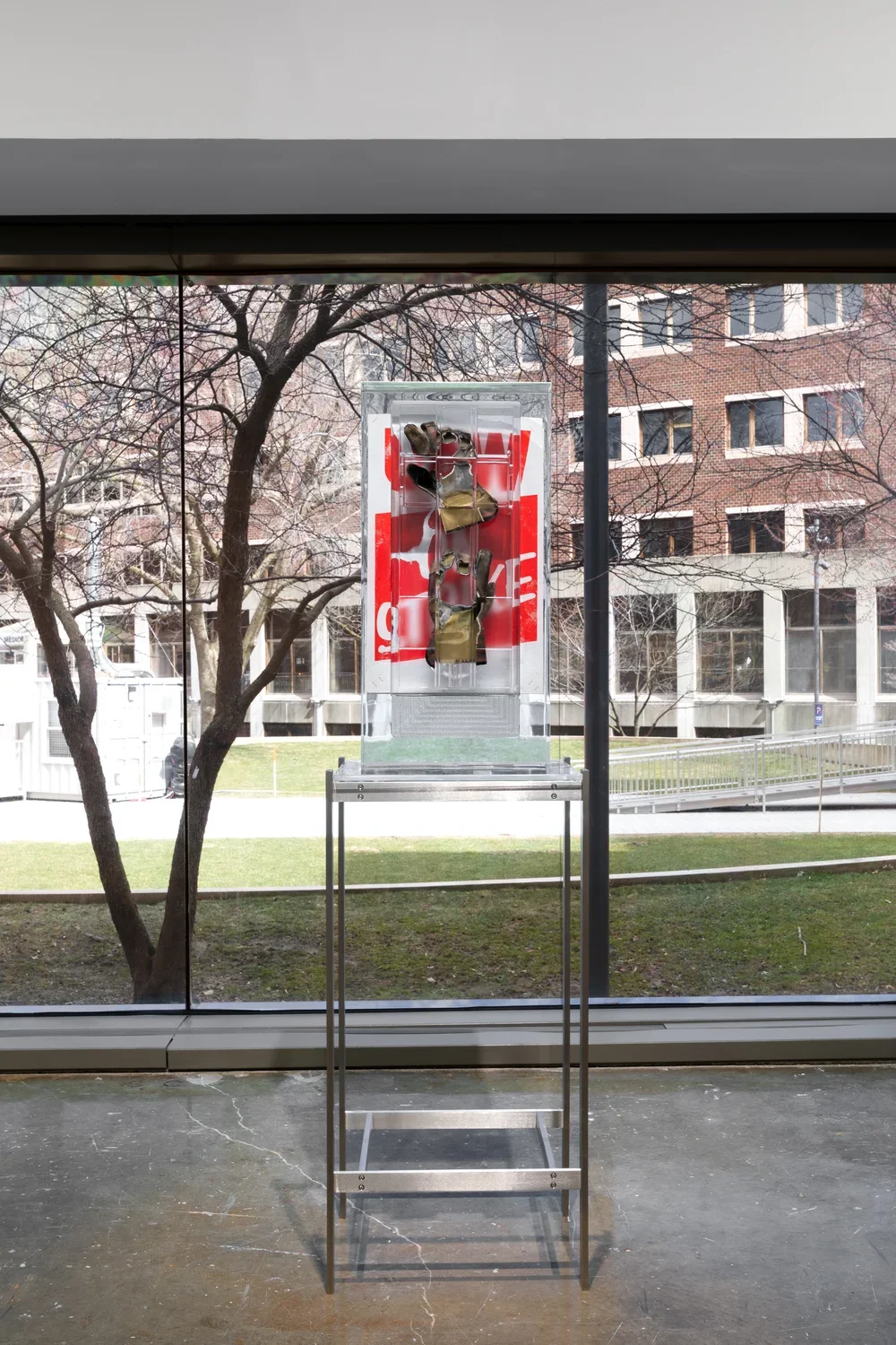 Installation view of a pair of work gloves and a red poster encased in a rectangular block of resin on a metal stand with a large window overlooking a courtyard in the background. 
