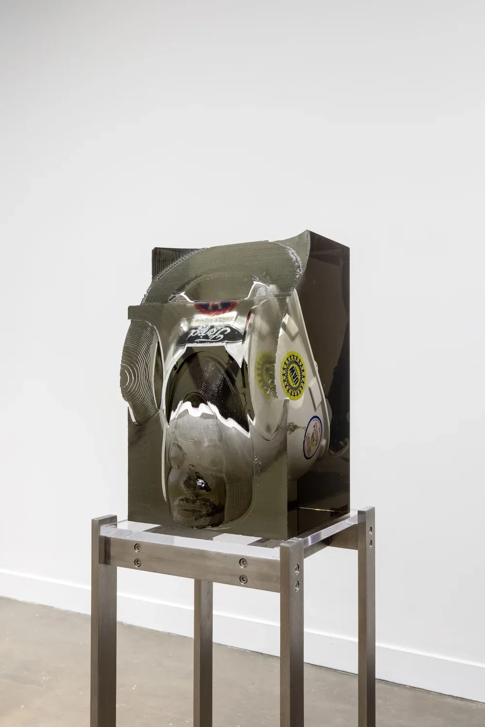 Installation view featuring a white Ford helmet with stickers on the side encased in a block of resin on a metal stand. 
