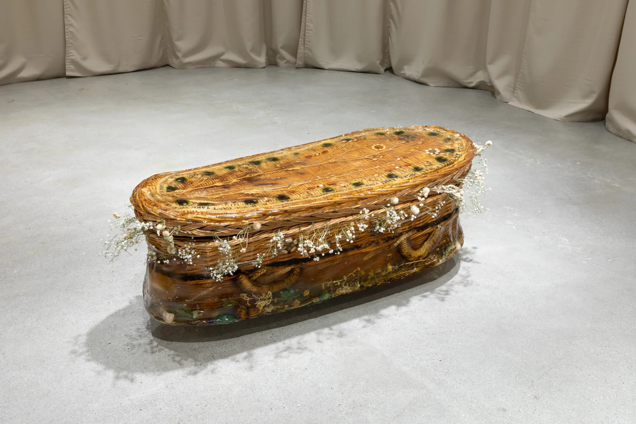 a rounded wicker casket encased in resinous gelatin sits  on the foor. Objects including syringes, pills, and beans adorn its top and lower sides, and dried flowers surround the opening between its lid and base. 