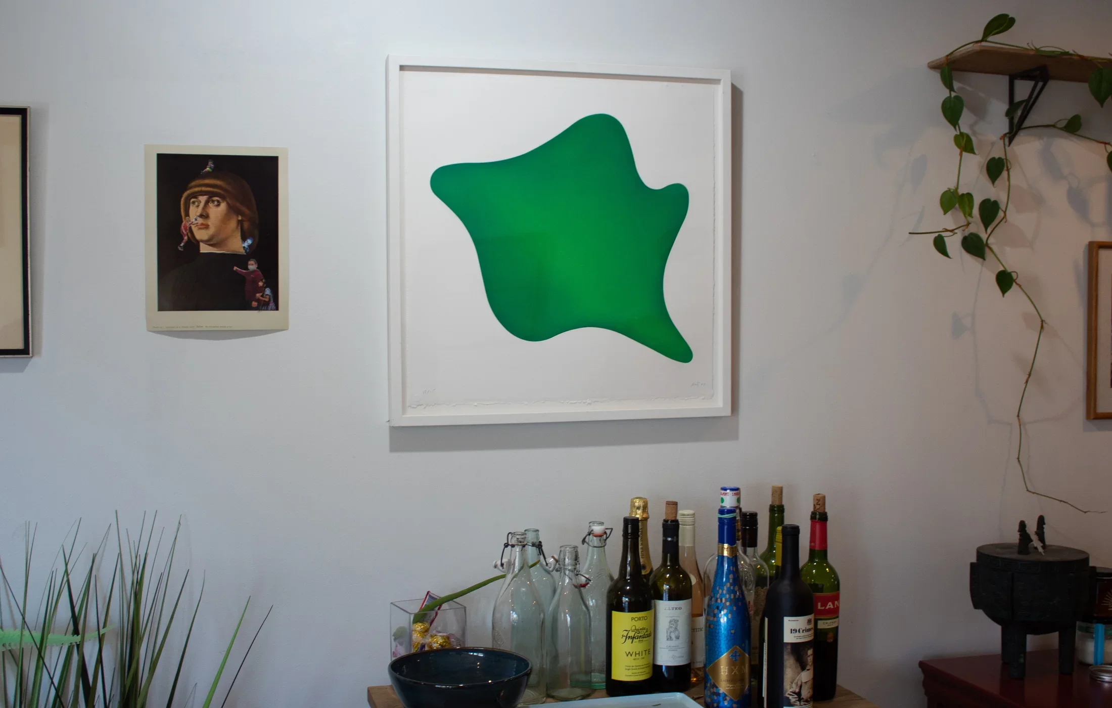 Photograph of a bar cart with alcohol on top, a green framed print above it and other artwork on both sides. 