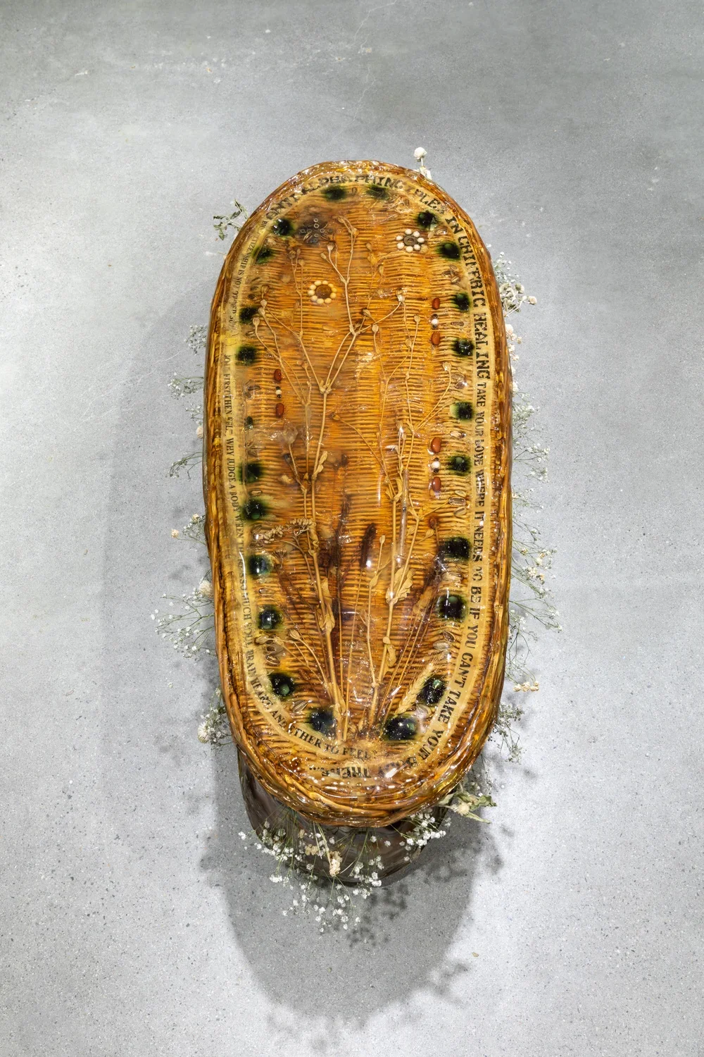 View from above of a wicker casket encased in resinous gelatin with objects including syringes, pills, and beans adorn on the top and dried flowers surround the opening between its lid and base.