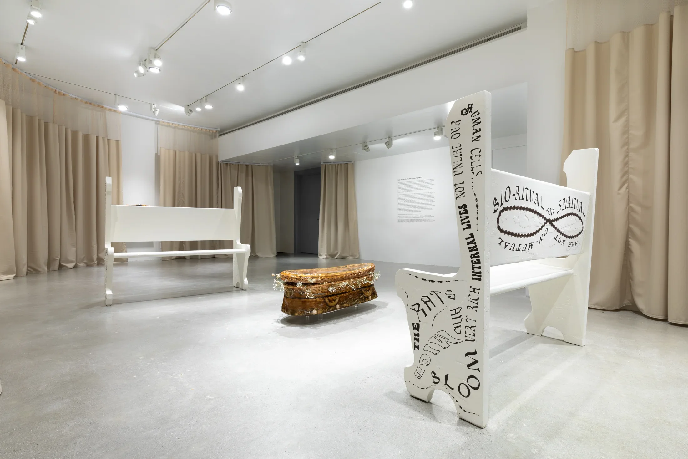 Two white church pews with text on sides and backs frame a sculpture of a wicker casket cast in gelatin with tan hospital curtains surrounding the interior of the gallery