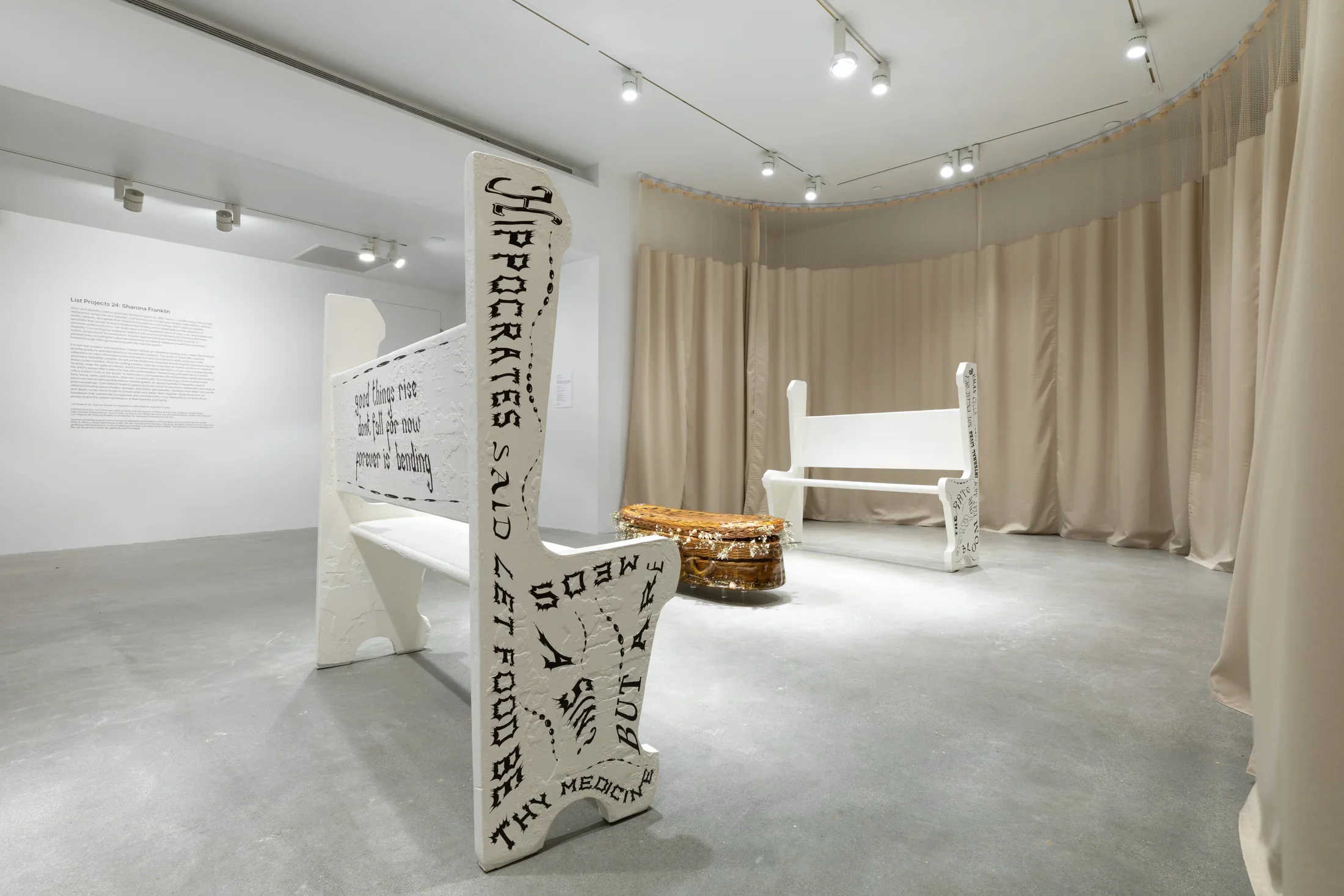 Sculpture of a willow wicker child size casket cast in gelatin sits on the floor in the middle of two white painted wooden church pews with black writing on the sides and back.