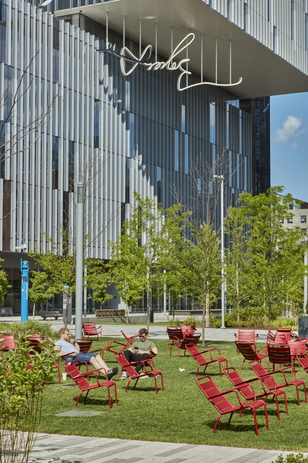 Lawn with red steel chairs in the foreground with a building in the back and newly installed LED sculpture of a hand drawn signature hanging down from the buildings overhand.