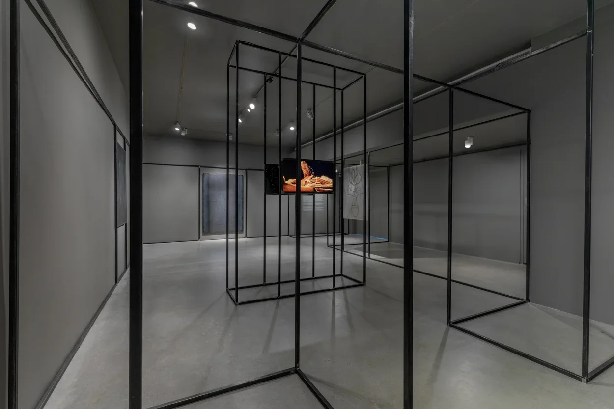 Gallery view with grey walls and cement flooring featuring a welded steel drawing on the back wall, a monitor in the center surrounded by black steel architecture that snakes along the periphery of the space. 