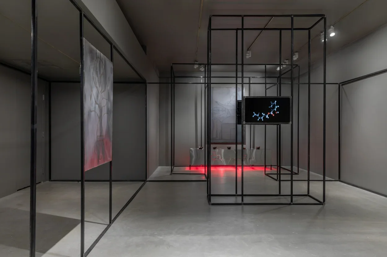 Gallery view with grey walls and cement flooring featuring welded steel drawings on the left and right, a monitor in the center, a steel platform at rear glows red from heat lamps underneath, and a black steel architecture snakes along the periphery of the space.  