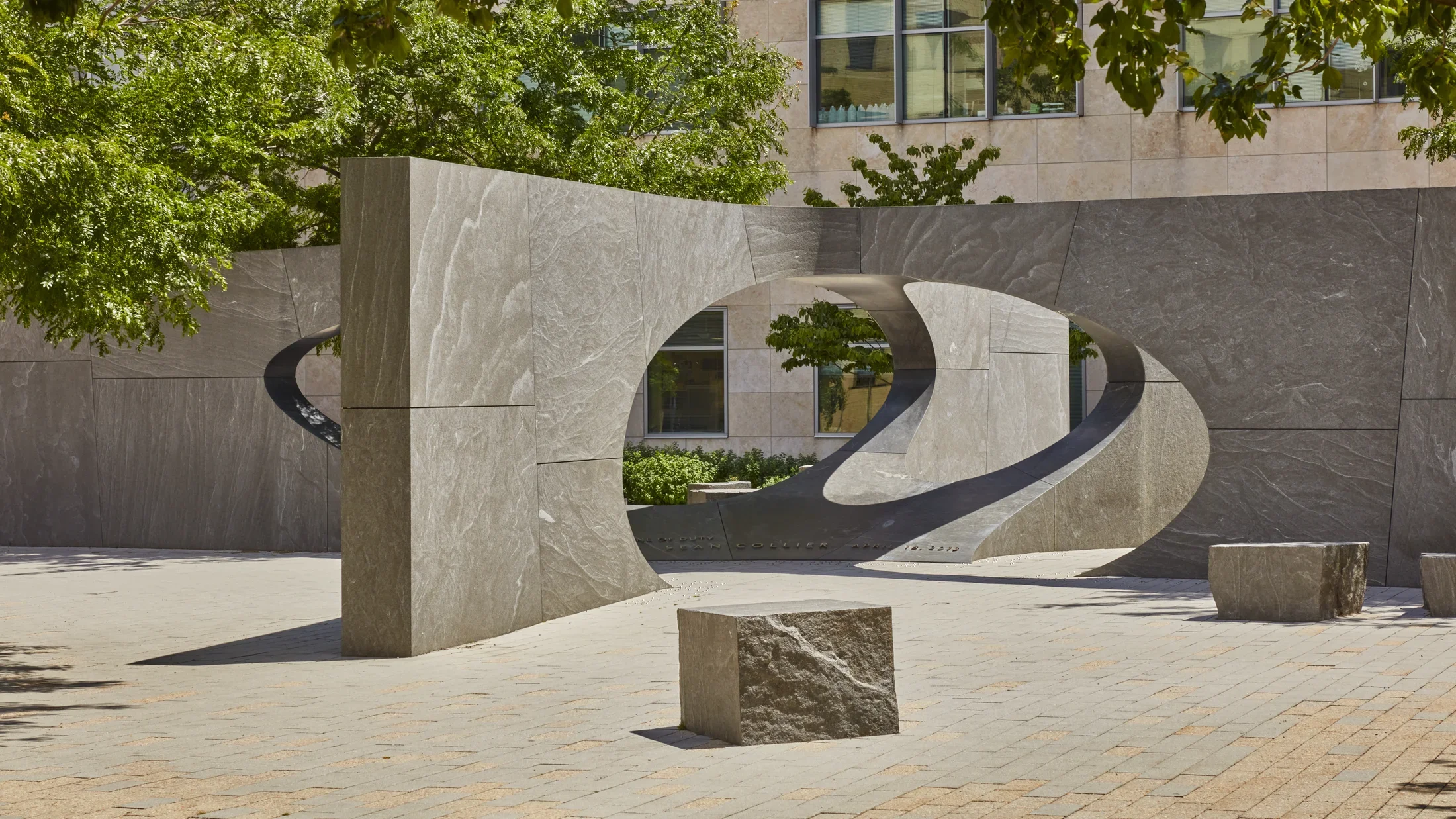 Granite open air sculpture featuring a rectangular shape with ovals cut out in the middle surrounded by trees peaking through the top edge of the photo. 
