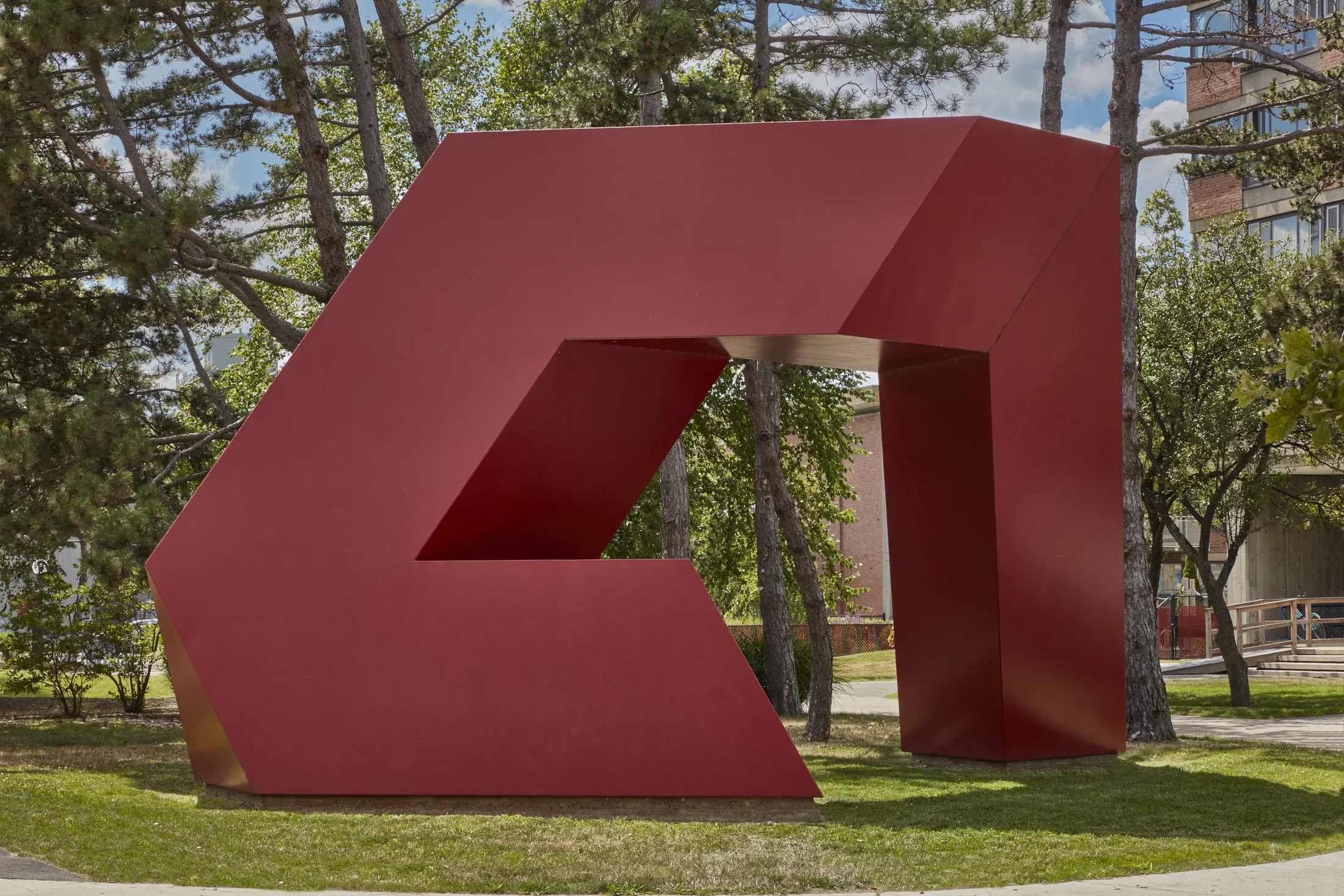 Red painted steel geometric sculpture with hard edges and rectangular shapes within on a small grass field. 