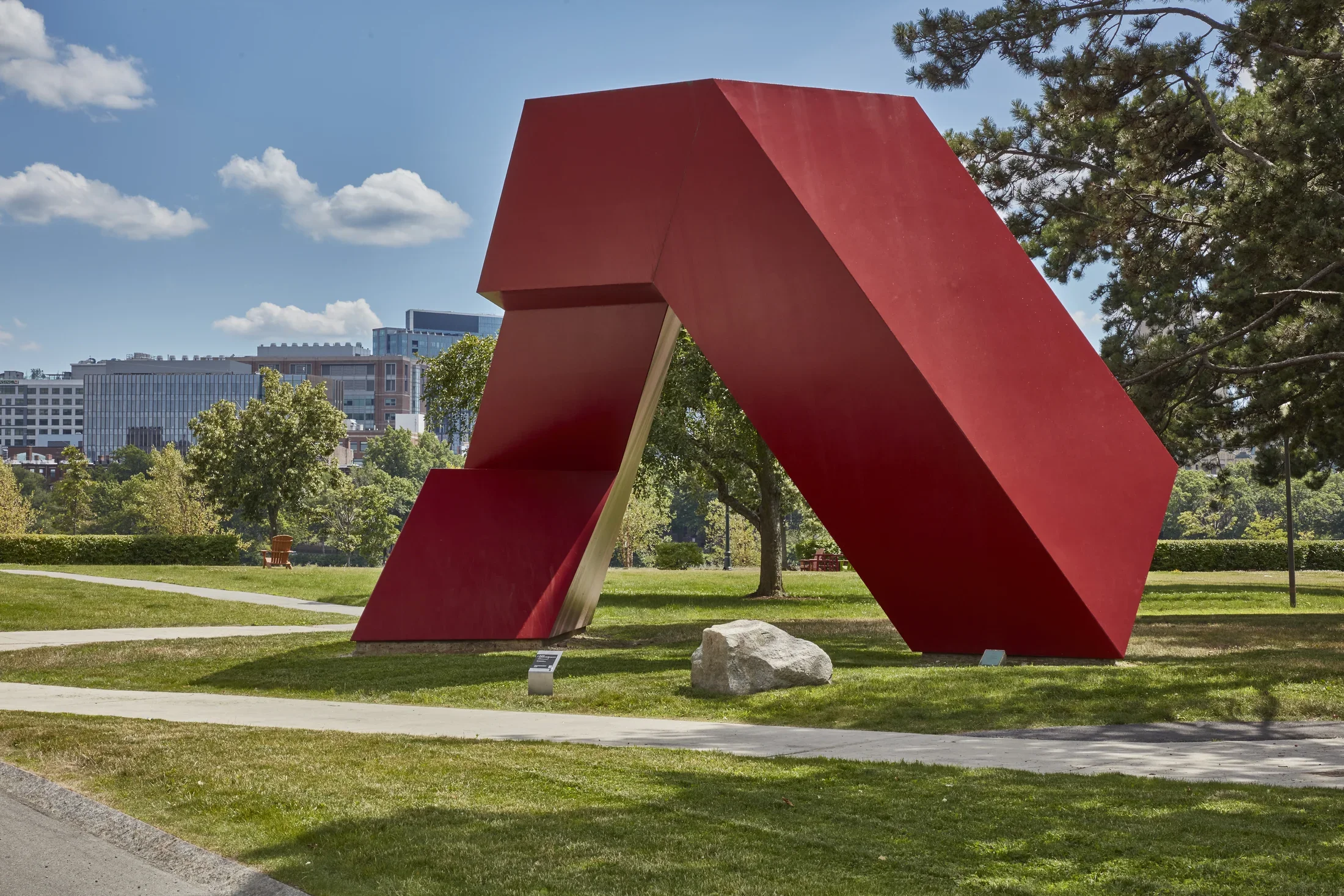 Red painted steel geometric sculpture with hard edges and rectangular shapes within on a grass field. 