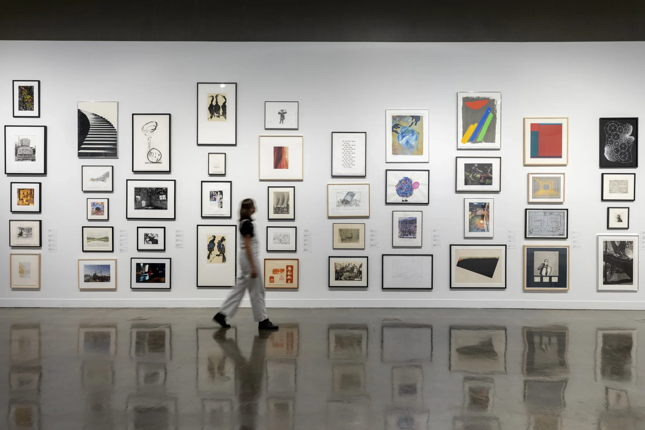 Installation view with framed artworks lining the wall and person in white overalls in motion walking through gallery.