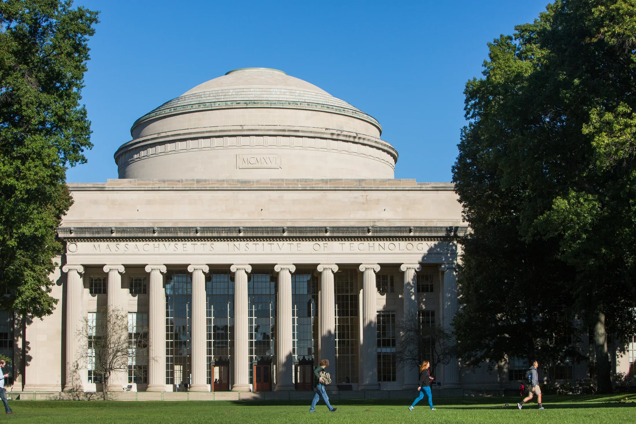 Domed building with tall columns and a blue sky, trees on each side, and a lawn in front with three students walking to the right. 