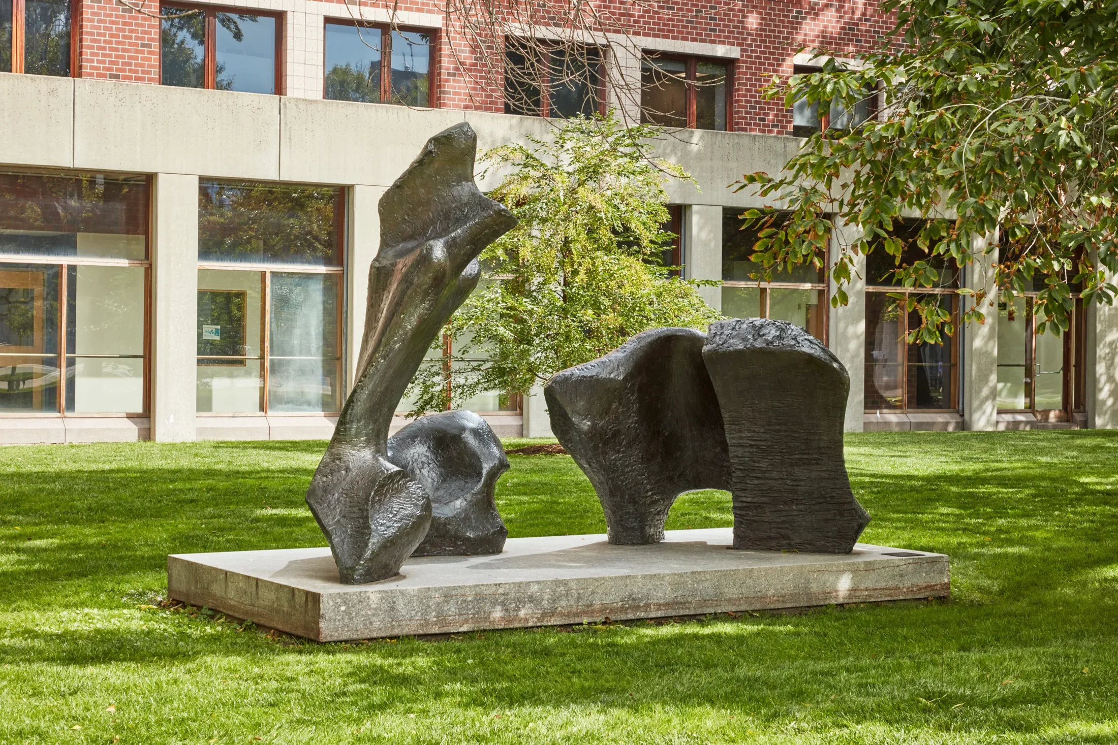Large scale abstract bronze sculpture on a stone pedestal in a field of grass with a brick building in the background. 