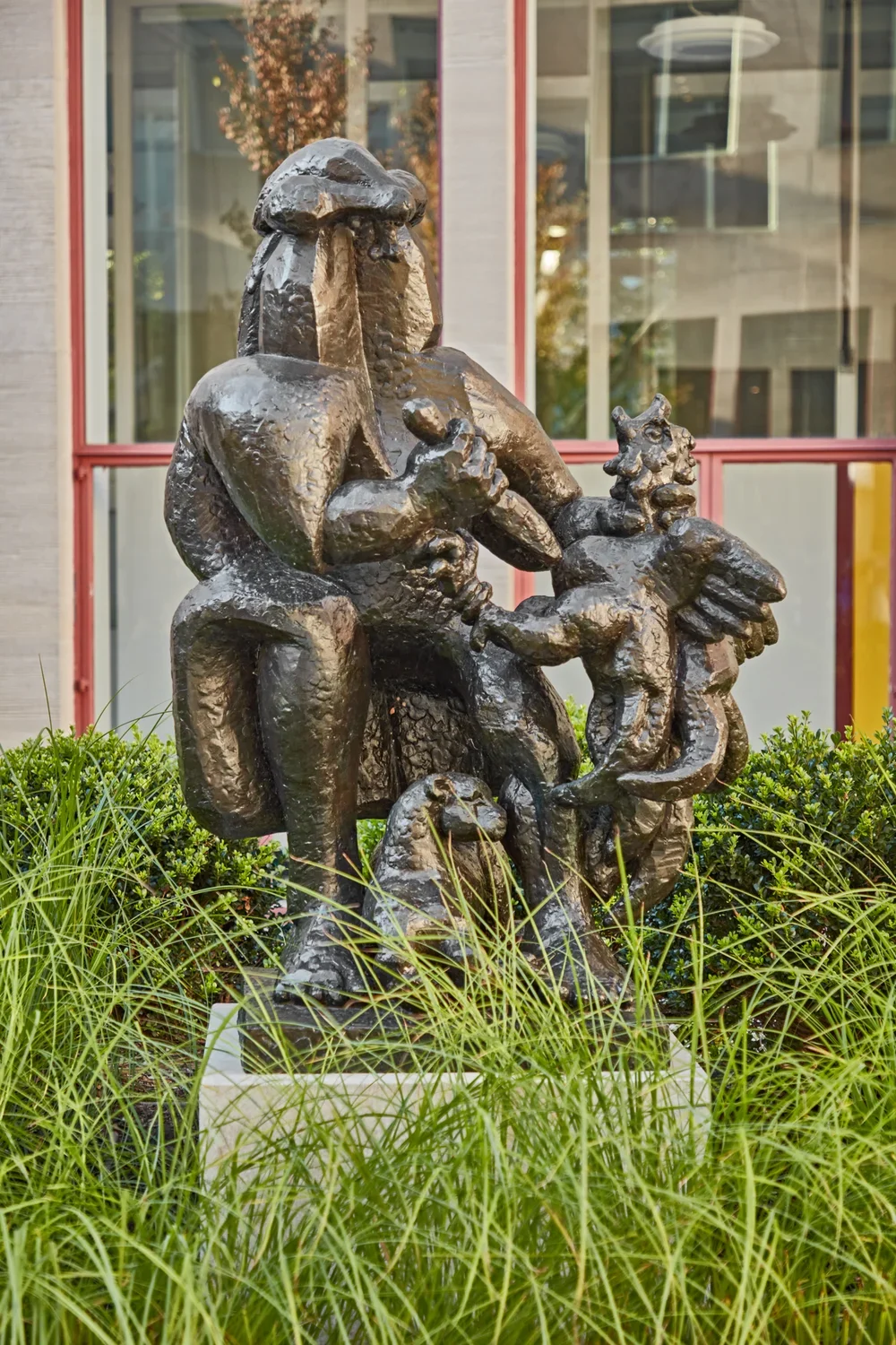 Bronze sculpture of an abstract figure on a stone pedestal in a grassy courtyard. 