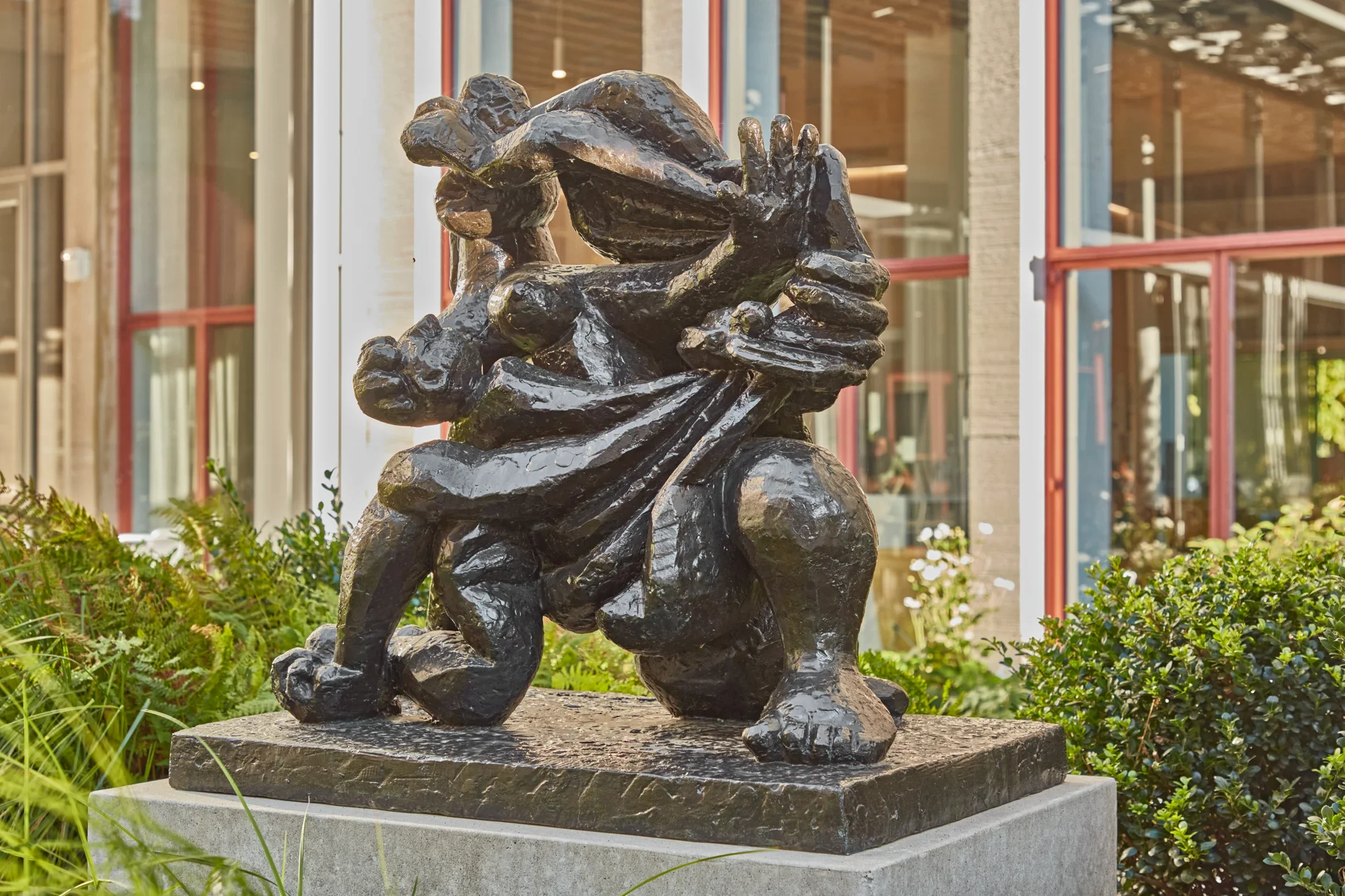 Bronze sculpture of abstract figures on a stone pedestal in a courtyard with greenery in the background. 