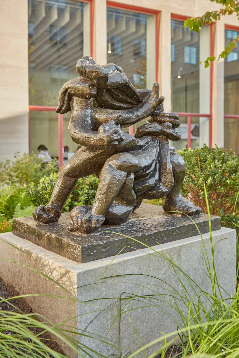 Bronze sculpture of abstract figures on a stone pedestal in a courtyard with greenery in the background. 