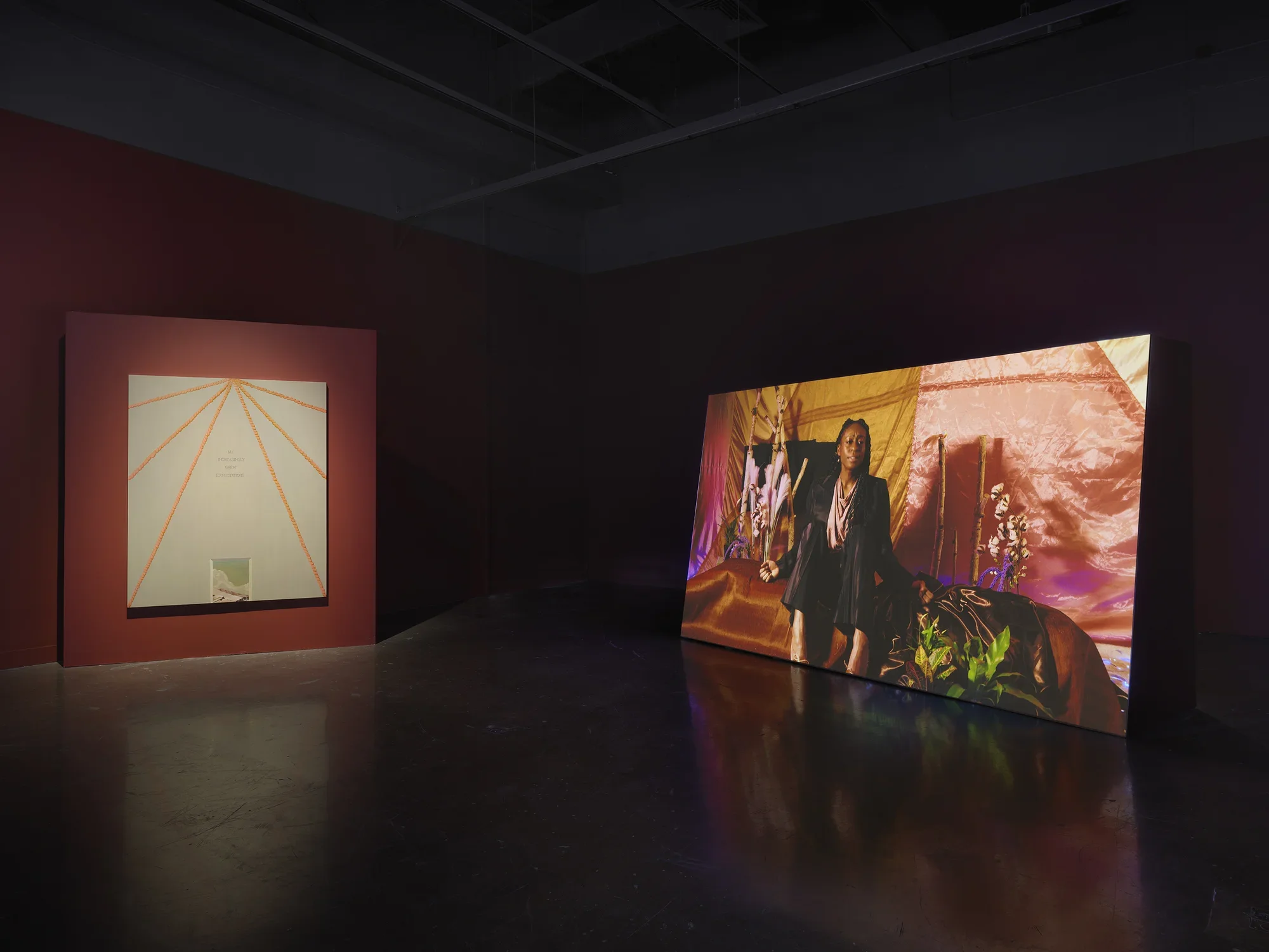 A beige and orange geometric painting is hung on a red wall nearby a large screen of a figure addressing the camera.