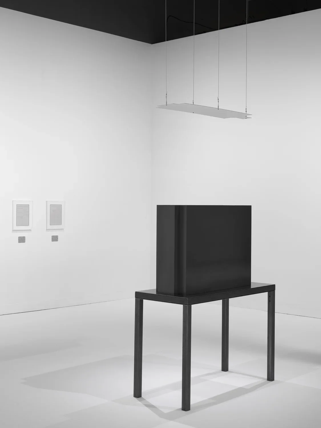 A dark steel table and geometric sculpture with a suspended mirror is opposite two framed works in a white walled and floored gallery.