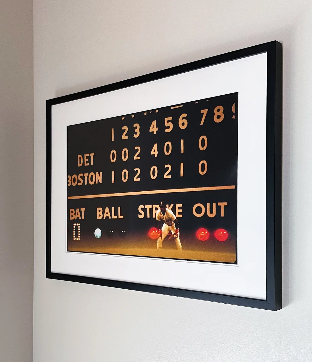 Black framed photograph of a baseball score board that reads "Bat Ball Strike Out". The photograph is hanging on a beige wall.