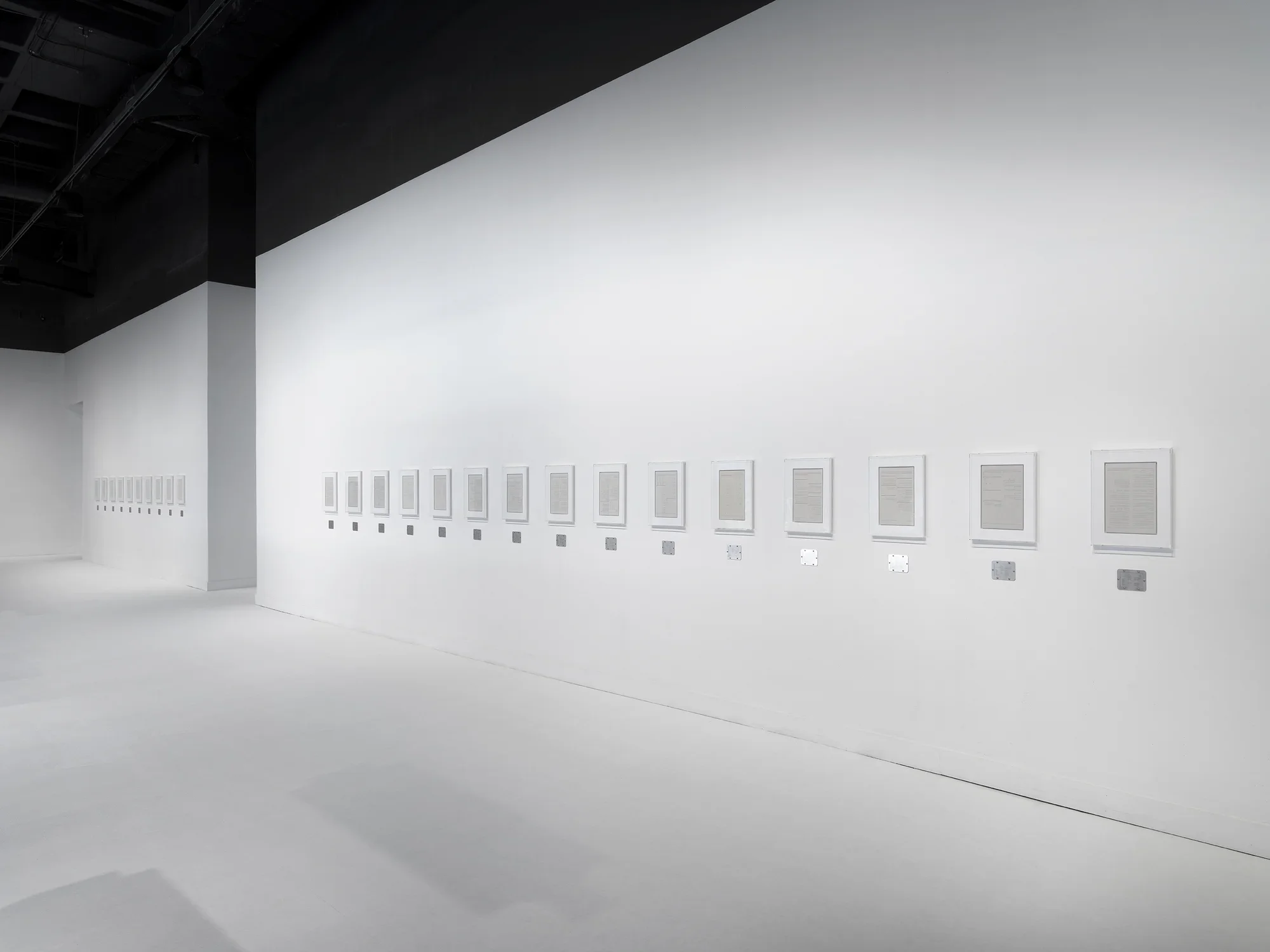 Several rectangular plaster works in white frames are hung horizontally across white gallery walls, each with a horizontal steel plaque below.