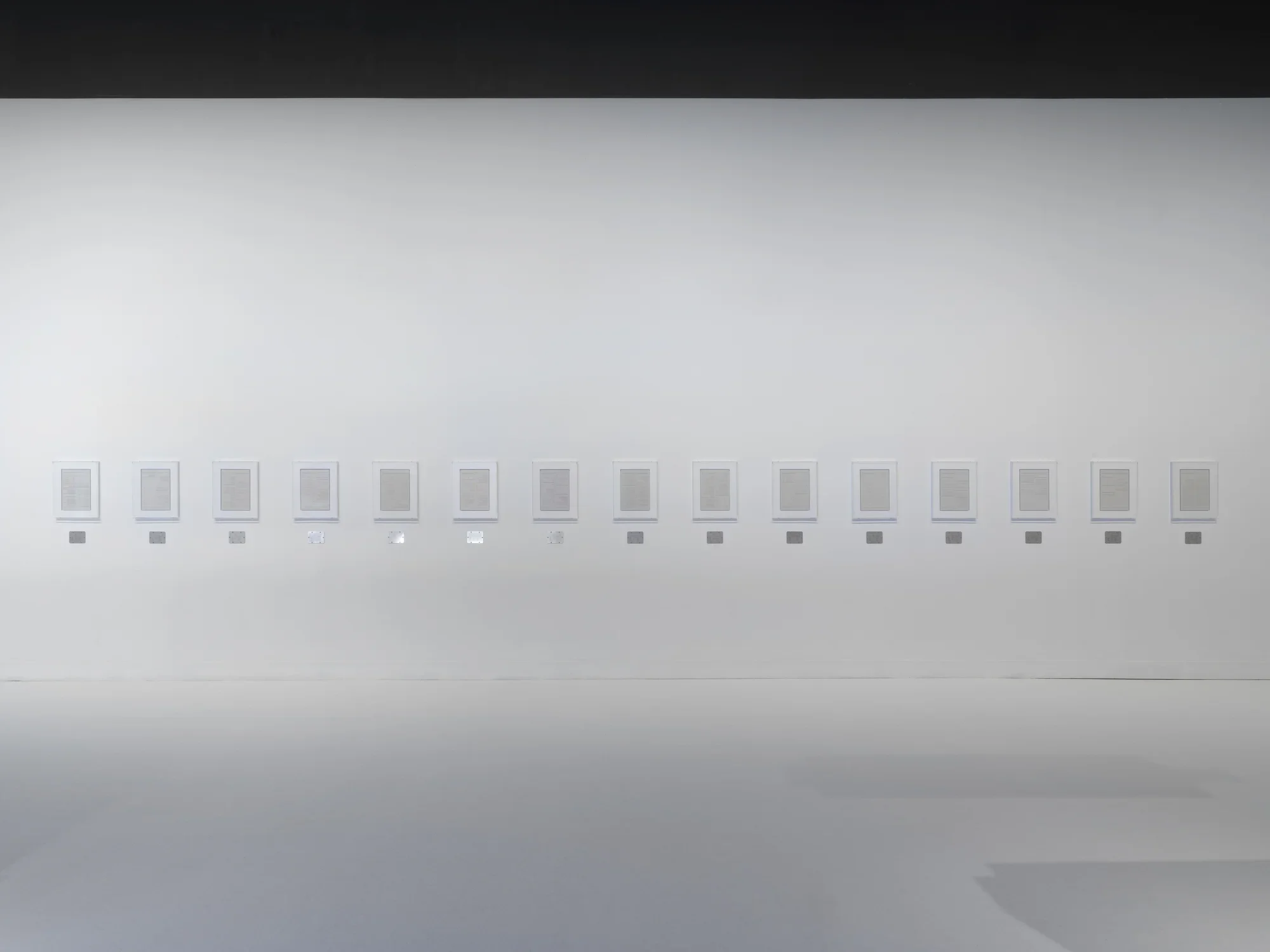 Fifteen page-sized plaster works in white frames are hung horizontally across white gallery walls, each with a small steel plaque below it.