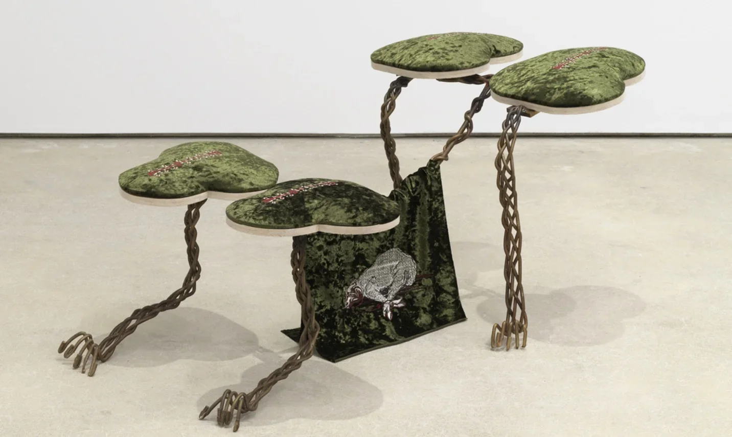 Four green crushed velvet panels each shaped like a trefoil spear to perch on top of a skeletal armature of sinewy braided bronze giving the overall impression of a four-legged animal crouching on its forelegs. At the animal’s belly, a flag of the same green cloth bears an embroidered design of a dog that appears to be either mid-gallop or hogtied.